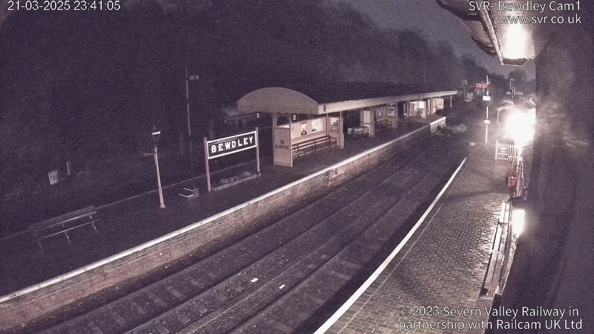 A nighttime view of a rain-slicked train station platform with a train partially visible in the distance.