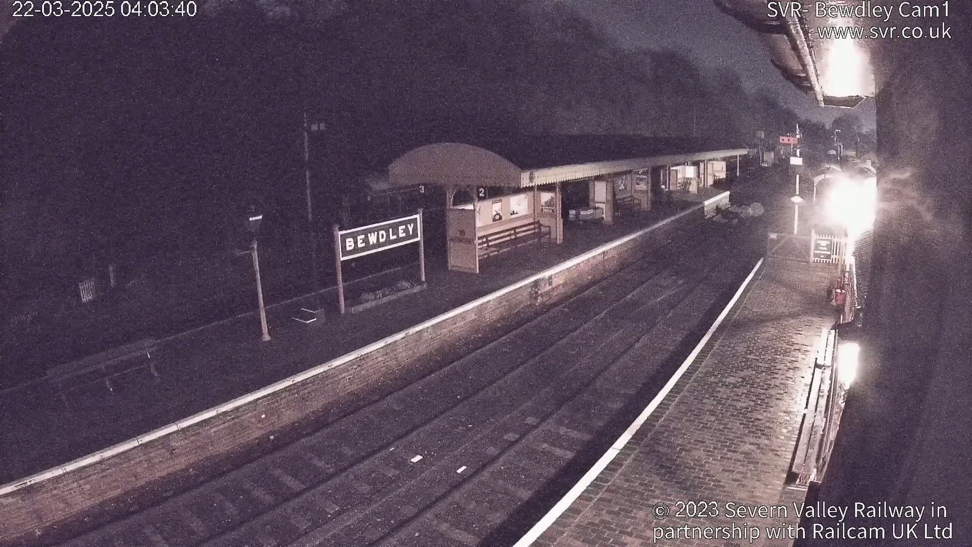 A nighttime view of a train arriving at a station platform with a "Bewdley" sign visible.