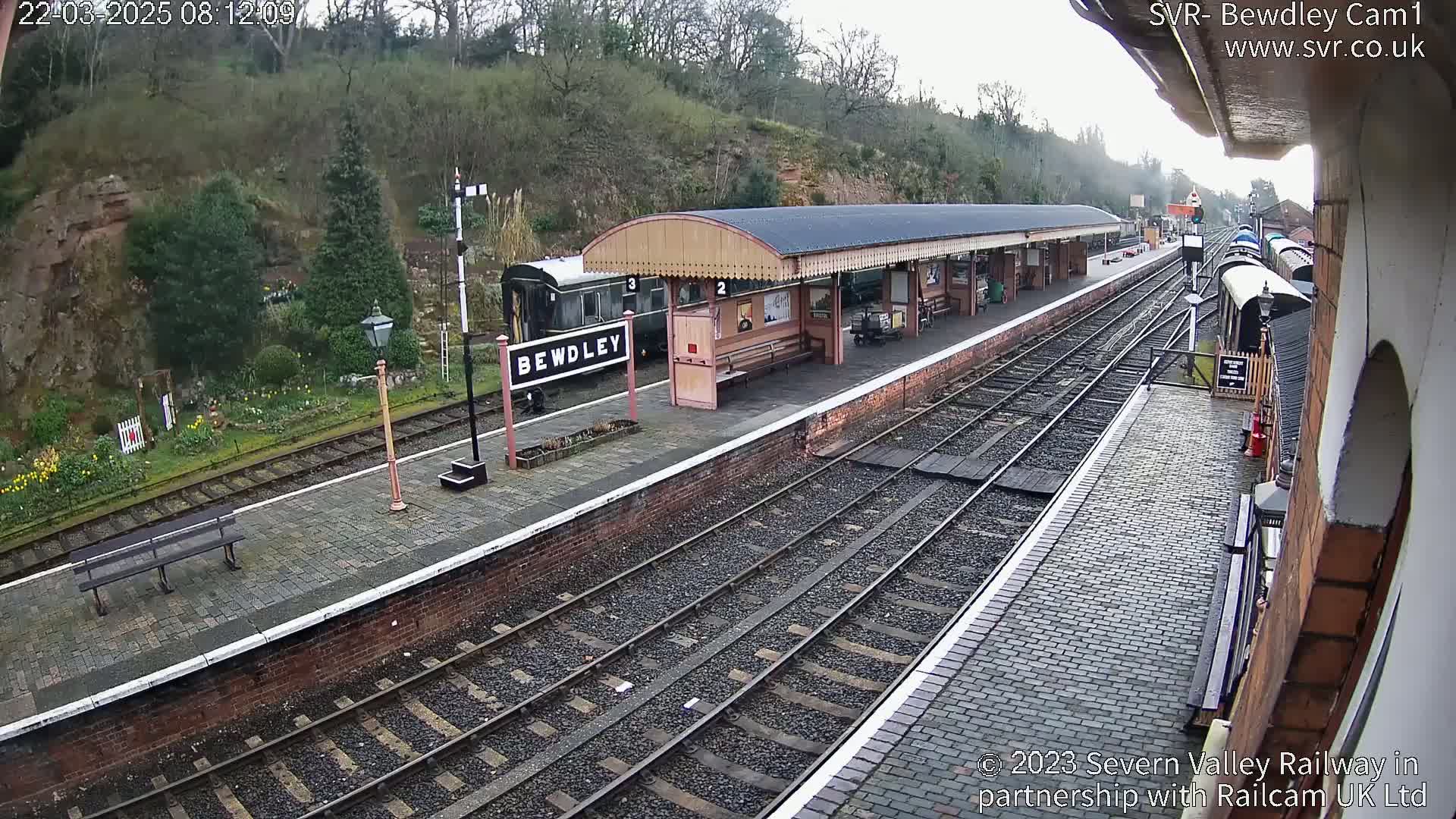 A train station platform with several train cars visible, situated on a hillside under an overcast sky.
