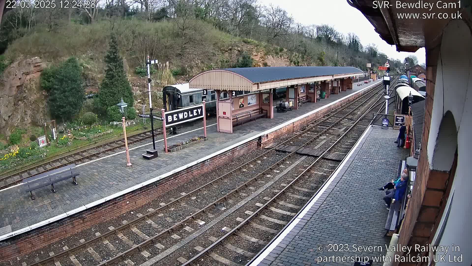 A train station platform with a covered waiting area, a railway track, and a partially visible train, under an overcast sky.