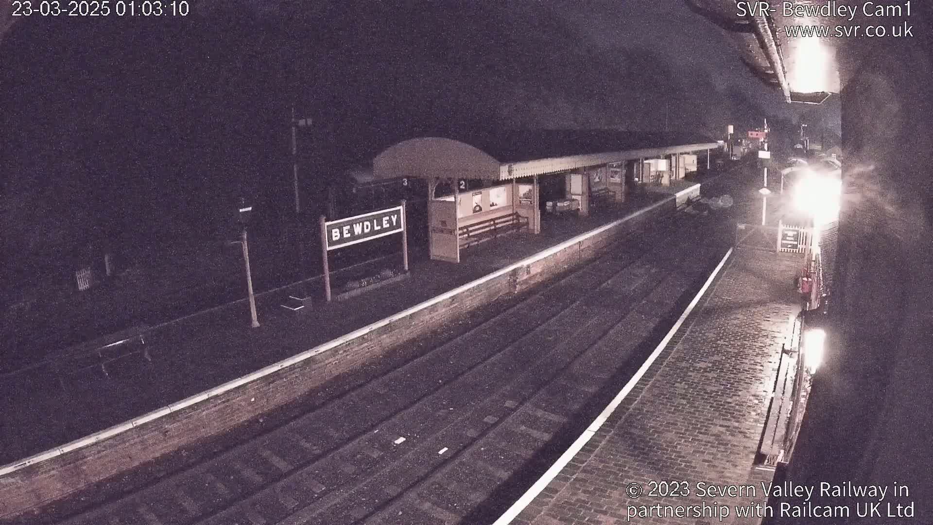 A nighttime view of a train arriving at a dimly lit train station with a cobblestone platform.