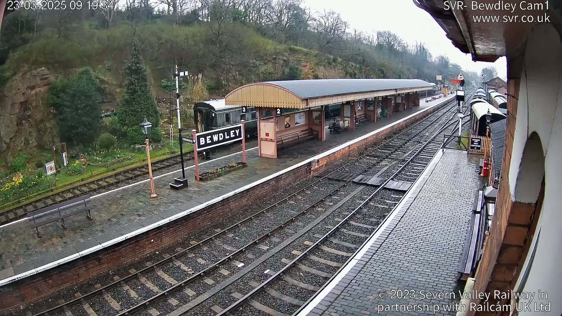 A wet train platform with several railway cars and a covered waiting area is seen on an overcast day.