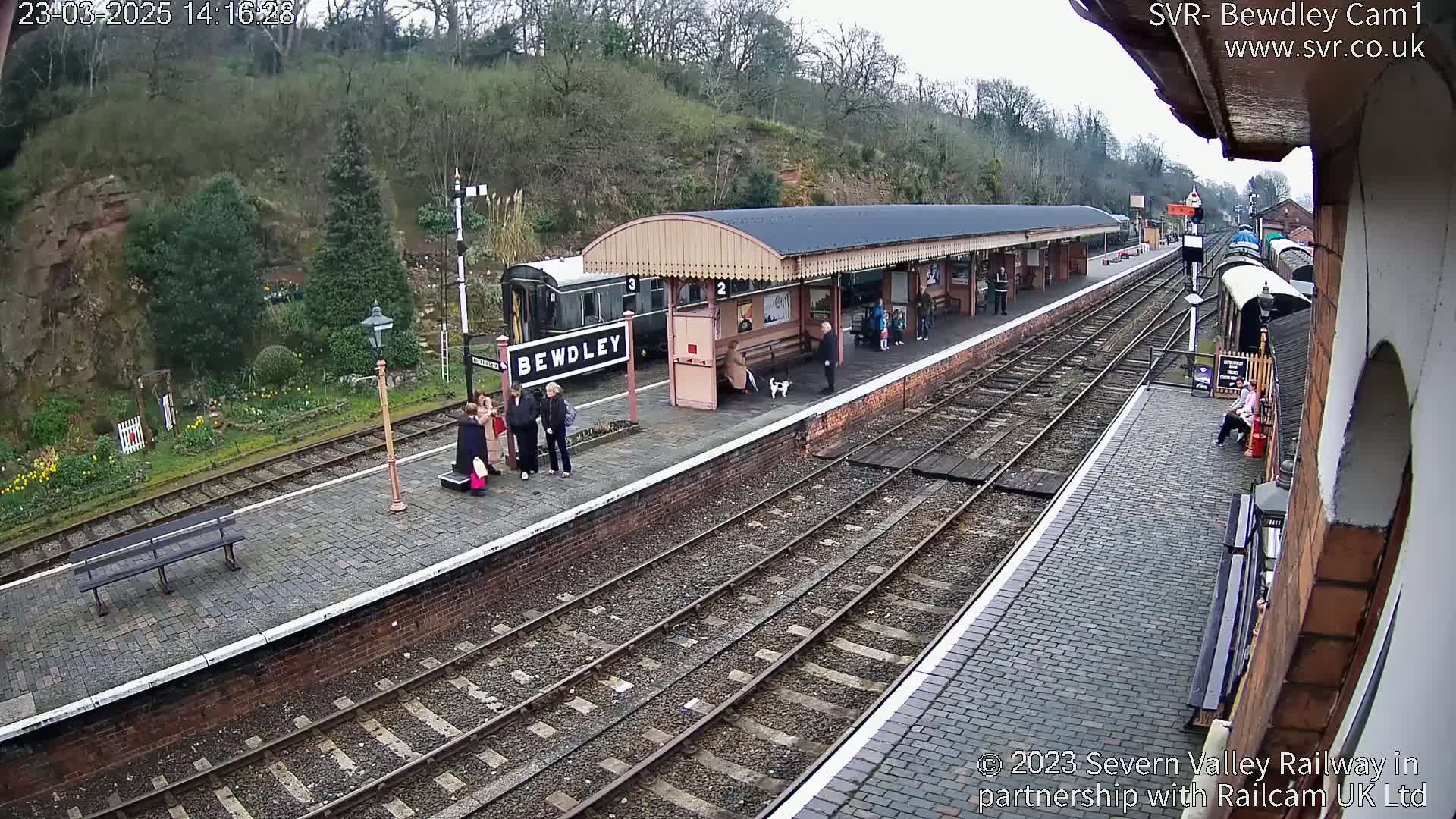 A train station platform with several people standing near a train and a covered waiting area, under an overcast sky.