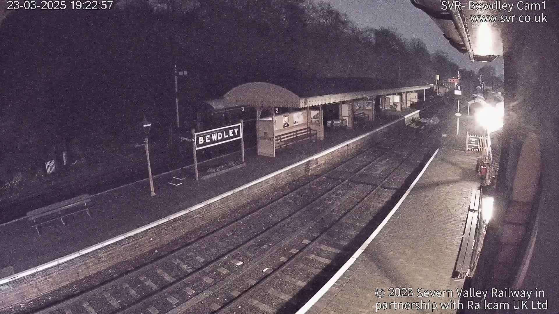 A nighttime view of a train station platform with train tracks and a partially visible train.