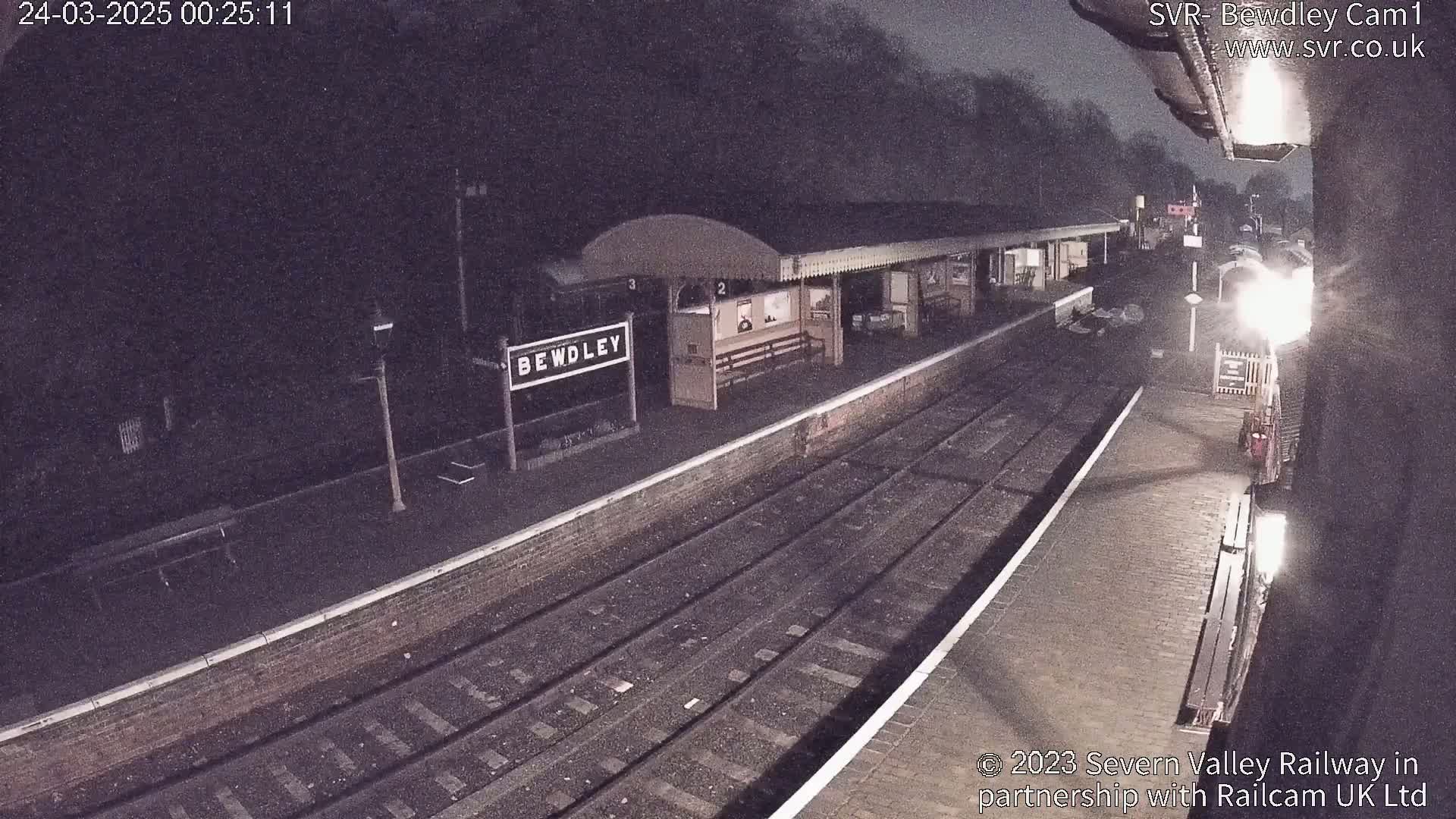 A nighttime view of a deserted train station platform with train tracks, a station building, and a sign indicating the station's name.