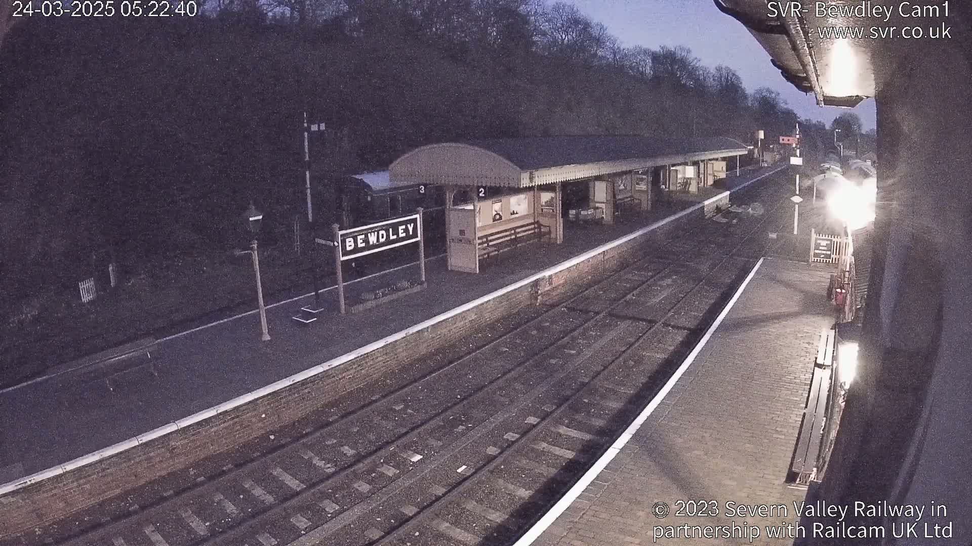A nighttime view of a train station platform with a train partially visible, under a dark sky.
