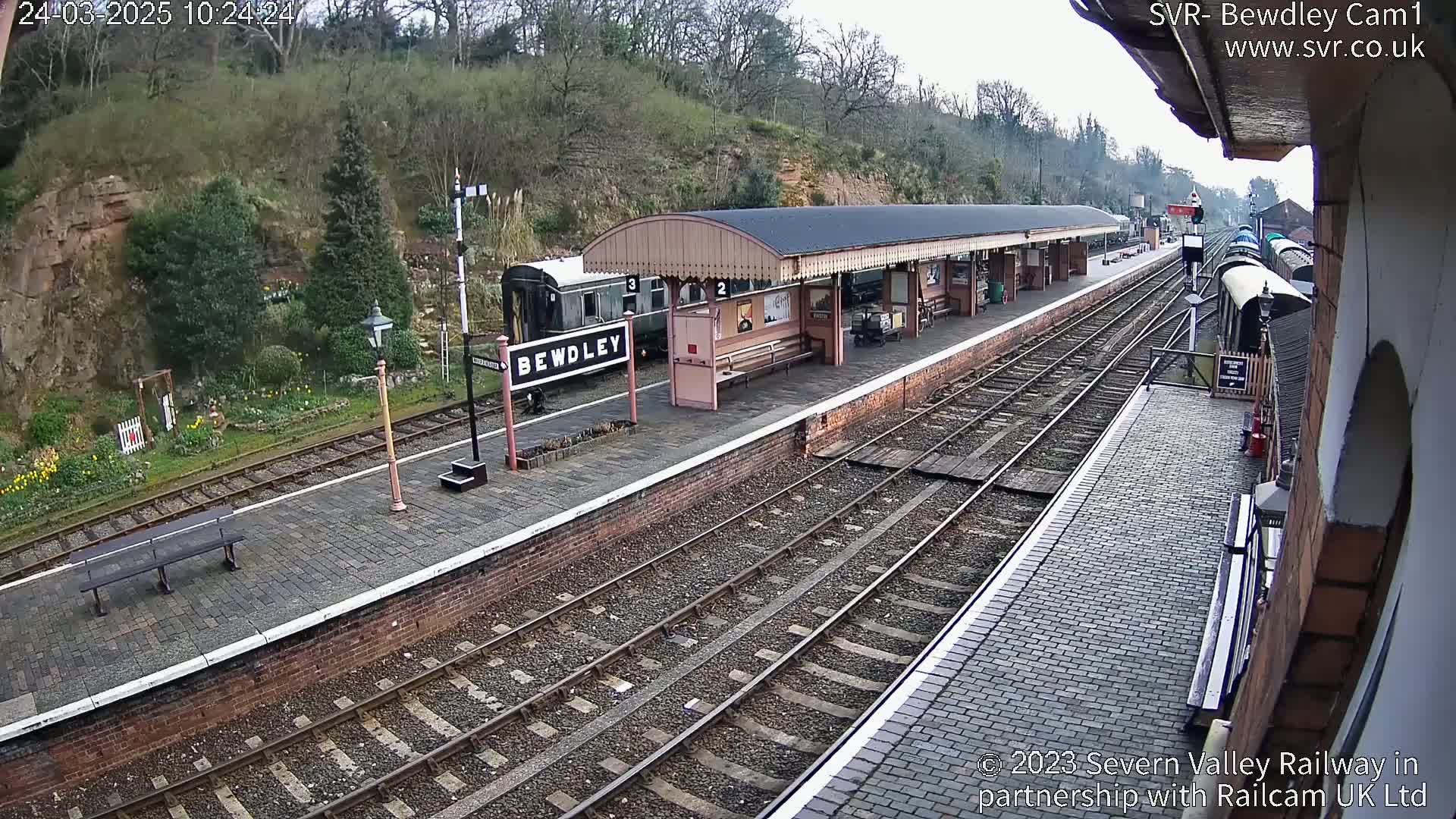 A train station platform with a covered waiting area, railway tracks, and a train partially visible, situated on a hillside with overcast skies.