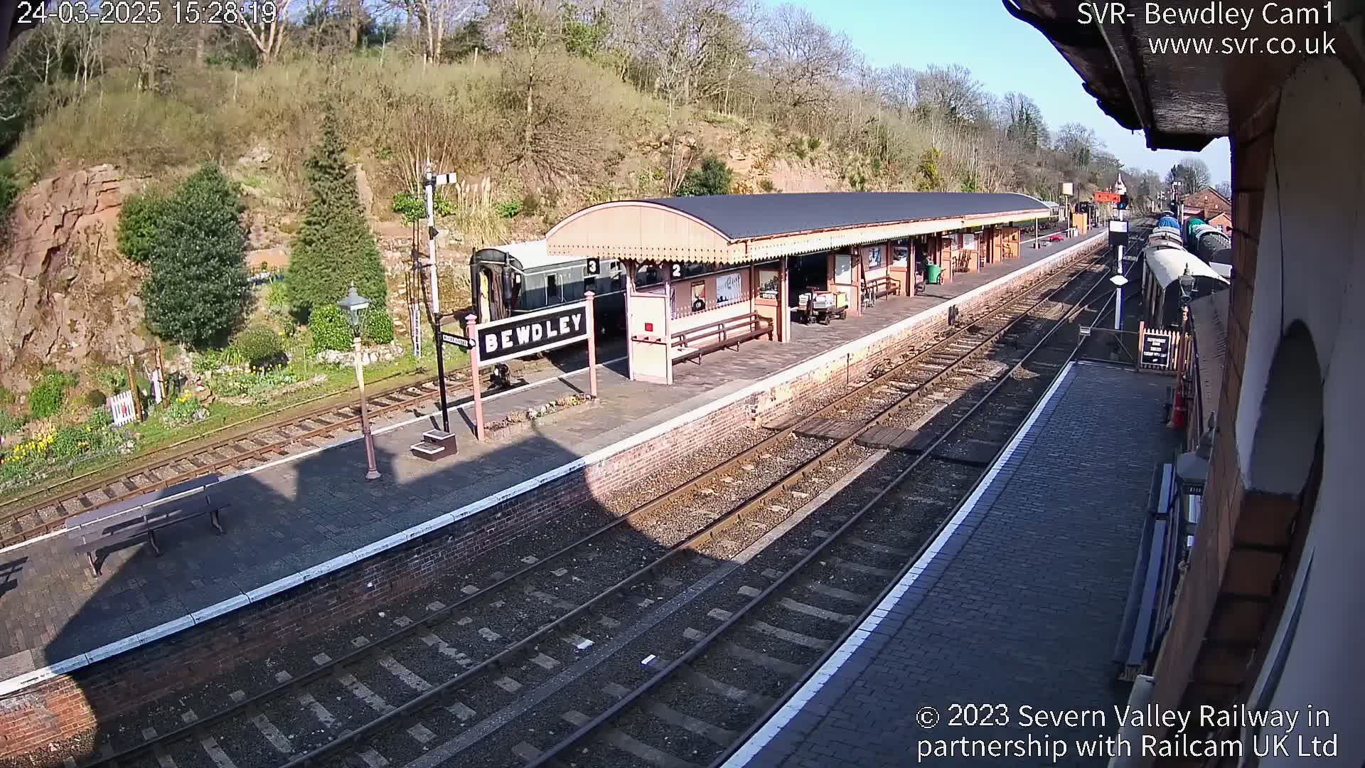 A train sits at a station platform on a sunny day, with a backdrop of lush green hills.