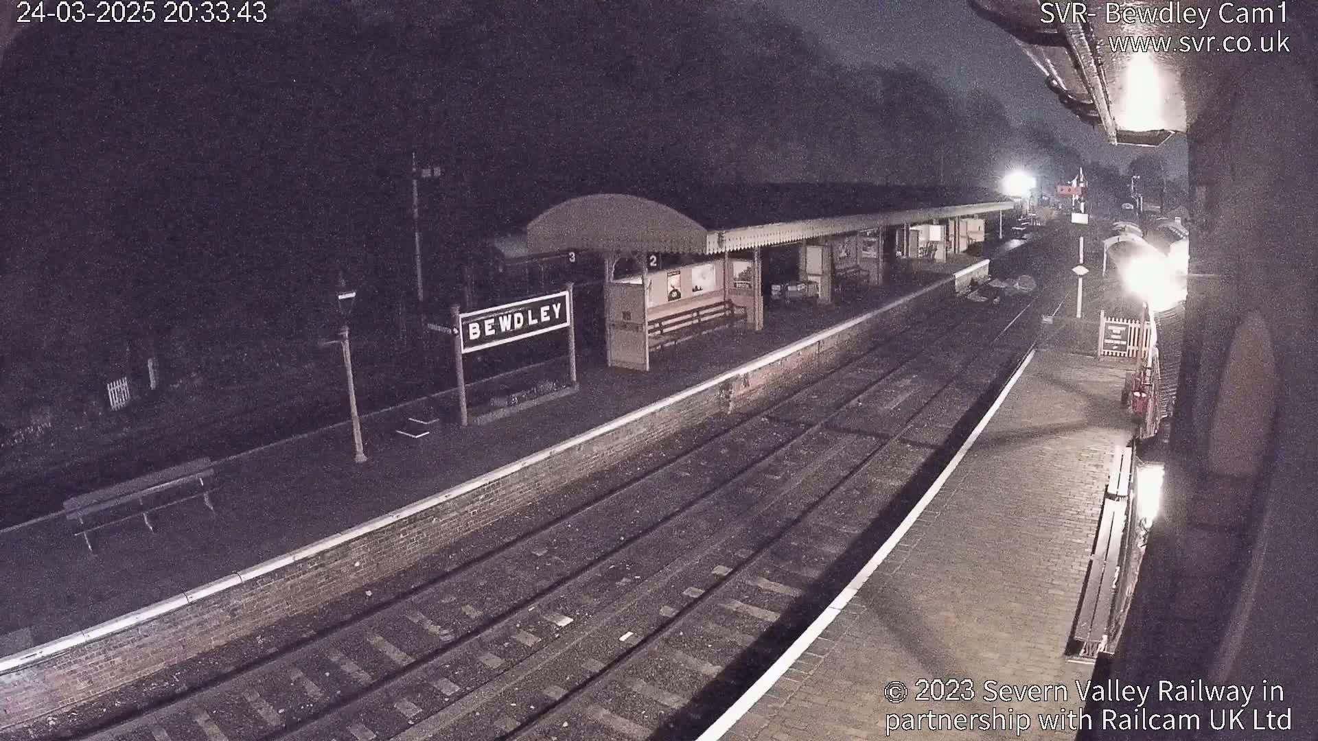 A nighttime view of a train station platform with train tracks, a station building, and a sign indicating "Bewdley," under a dark, possibly misty sky.