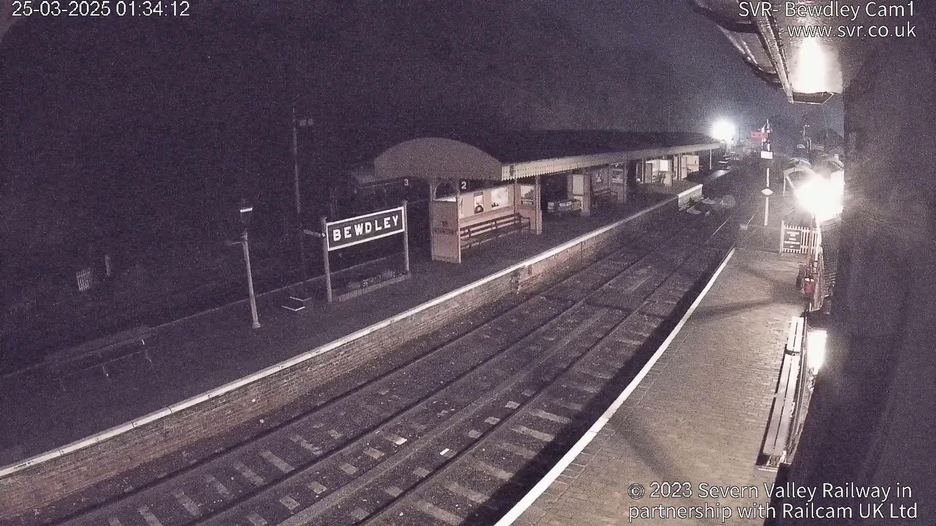 A nighttime view of a deserted train platform with several tracks and a partially visible train.