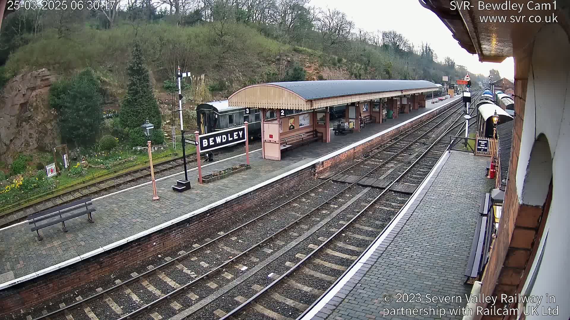 A train station platform with a covered waiting area, railway tracks, and a passenger train is visible on an overcast day.