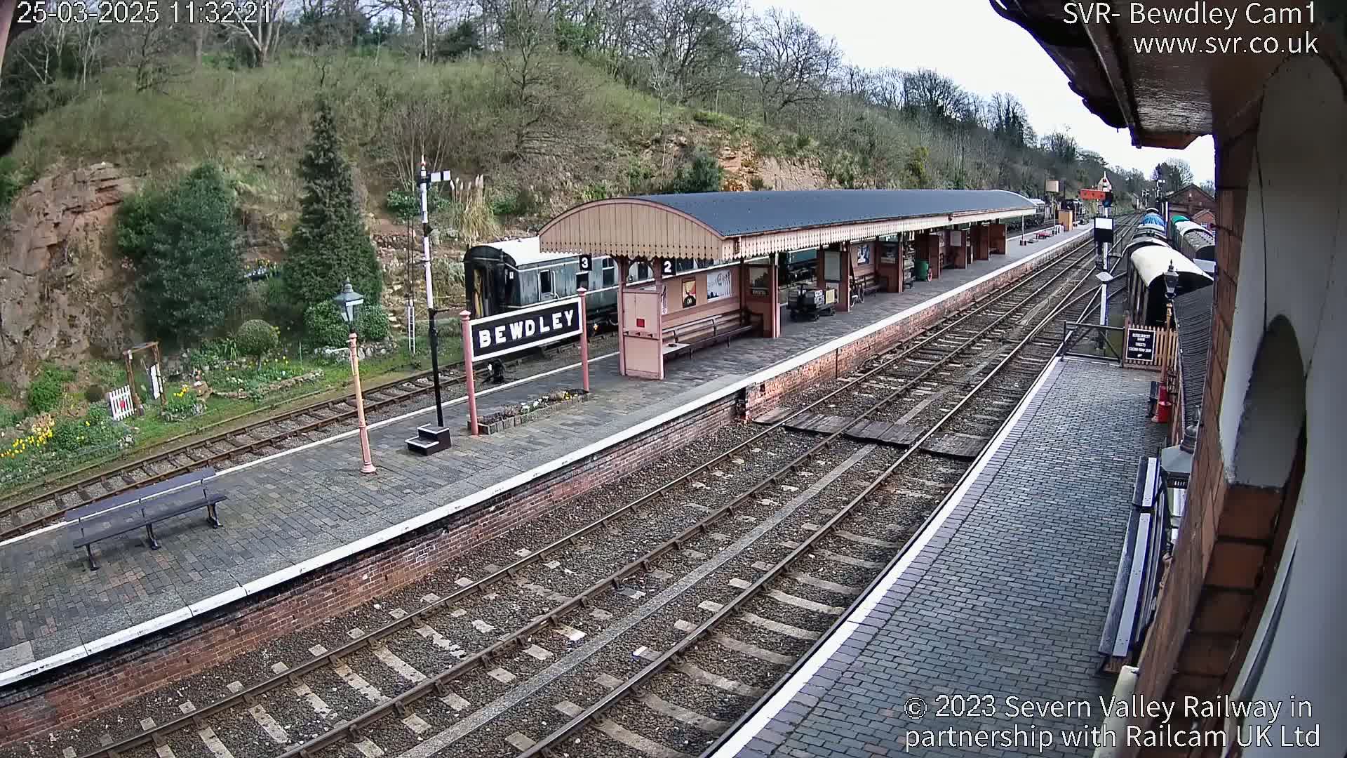 A train station platform with a railway track and a partially visible train under a partly cloudy sky.