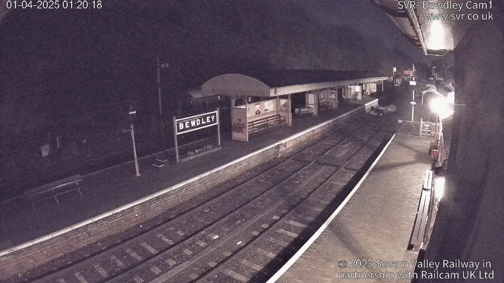 A nighttime view of a train station platform with train tracks and a partially visible train.