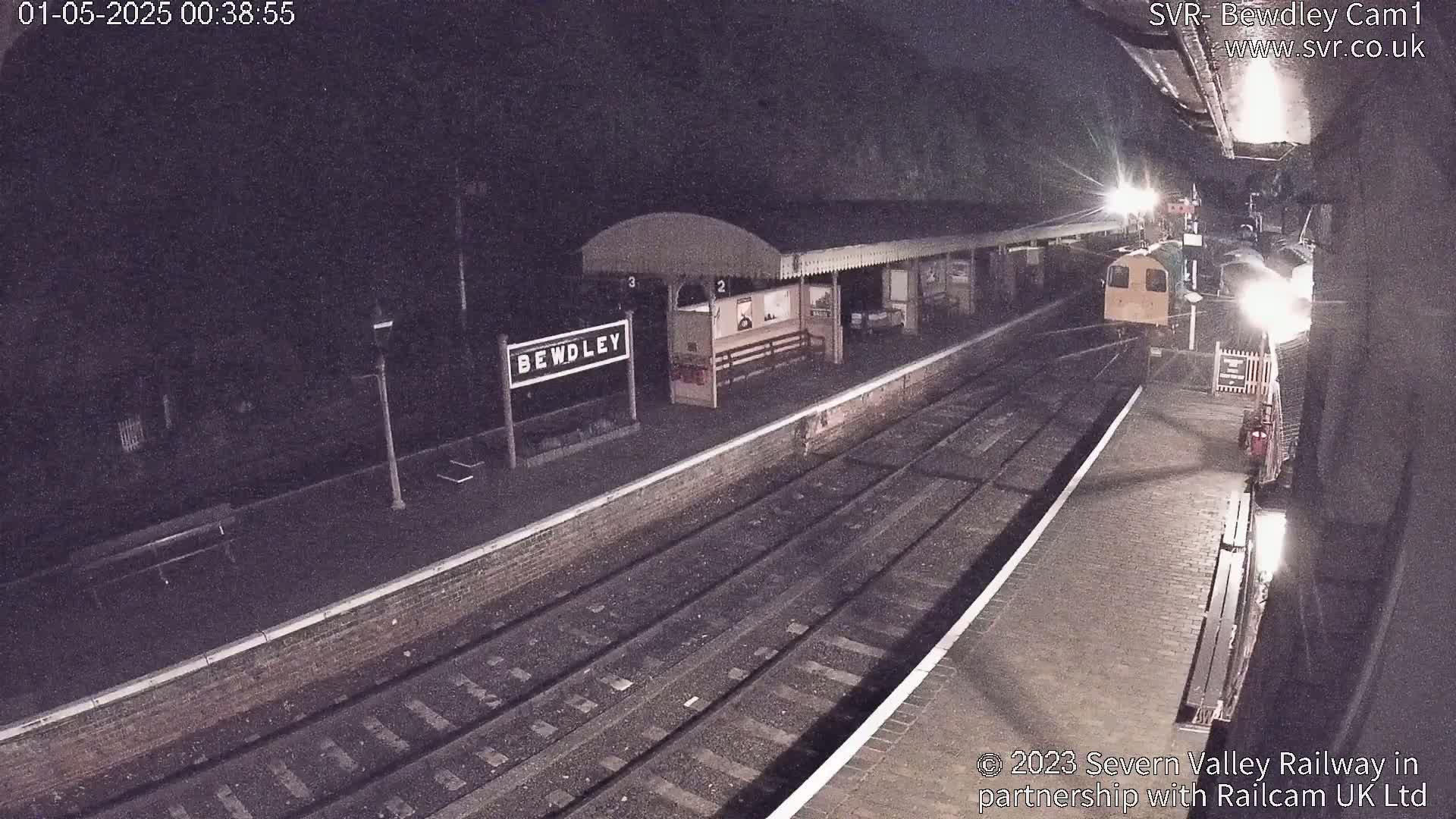 A nighttime view of a train station platform with a train partially visible under a dark sky.