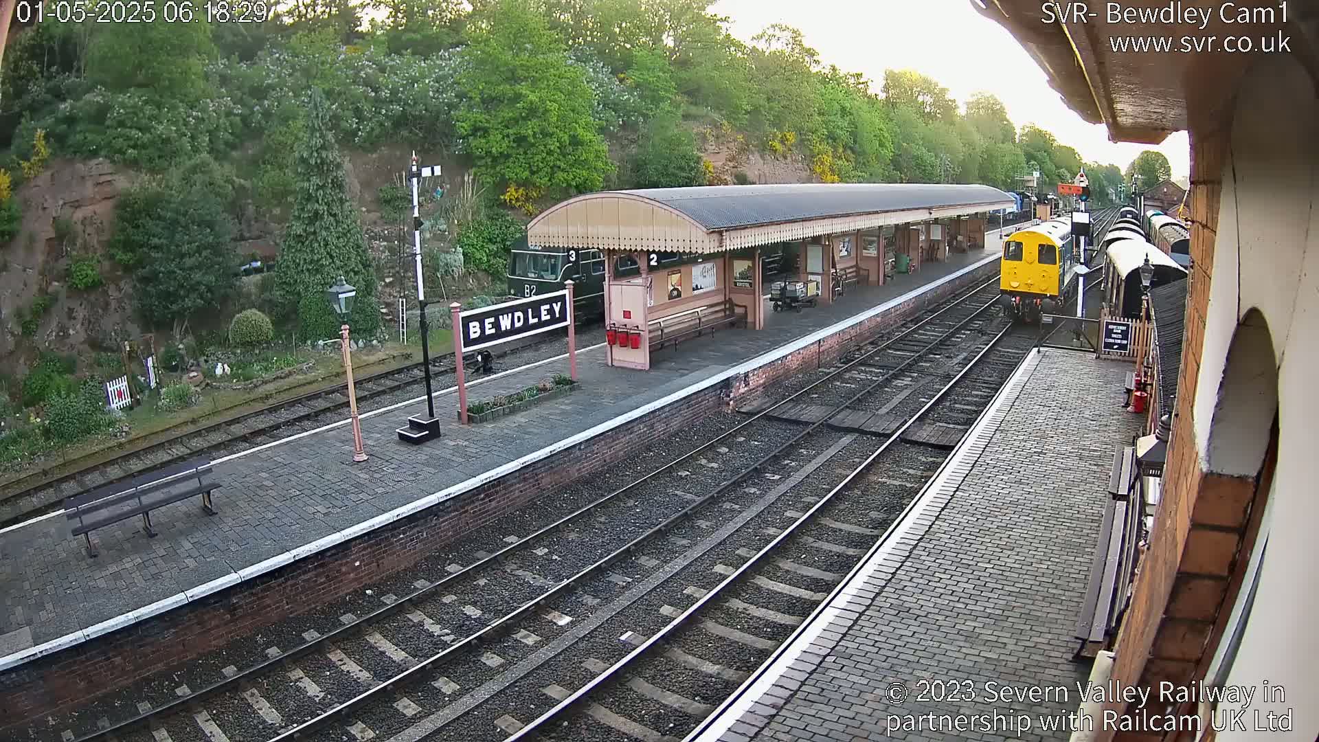 A train pulls into a railway station platform on a partly sunny morning, next to a brick building and lush green vegetation.