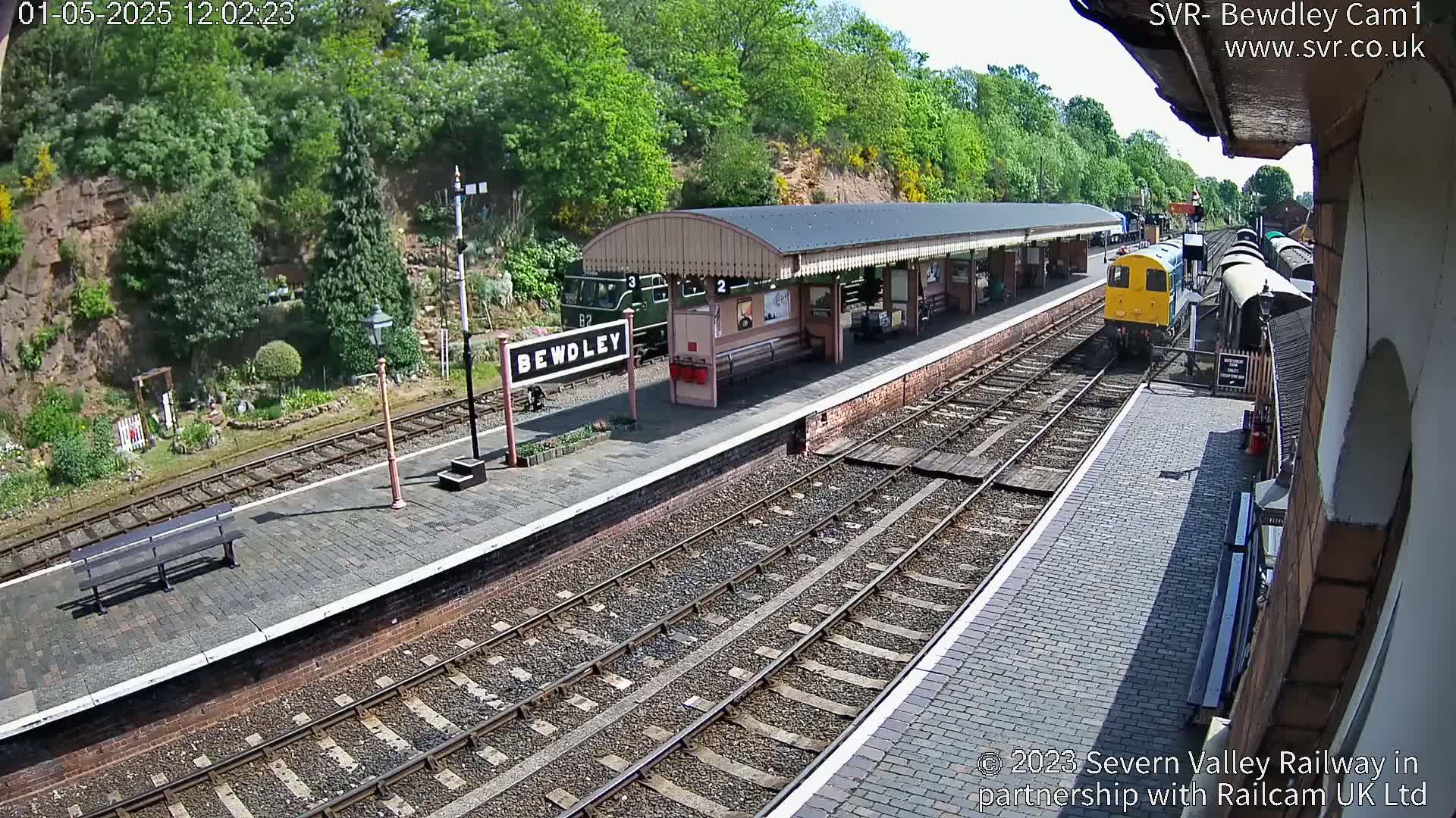 A train arrives at a railway station on a sunny day, passing a covered platform with a train already parked.