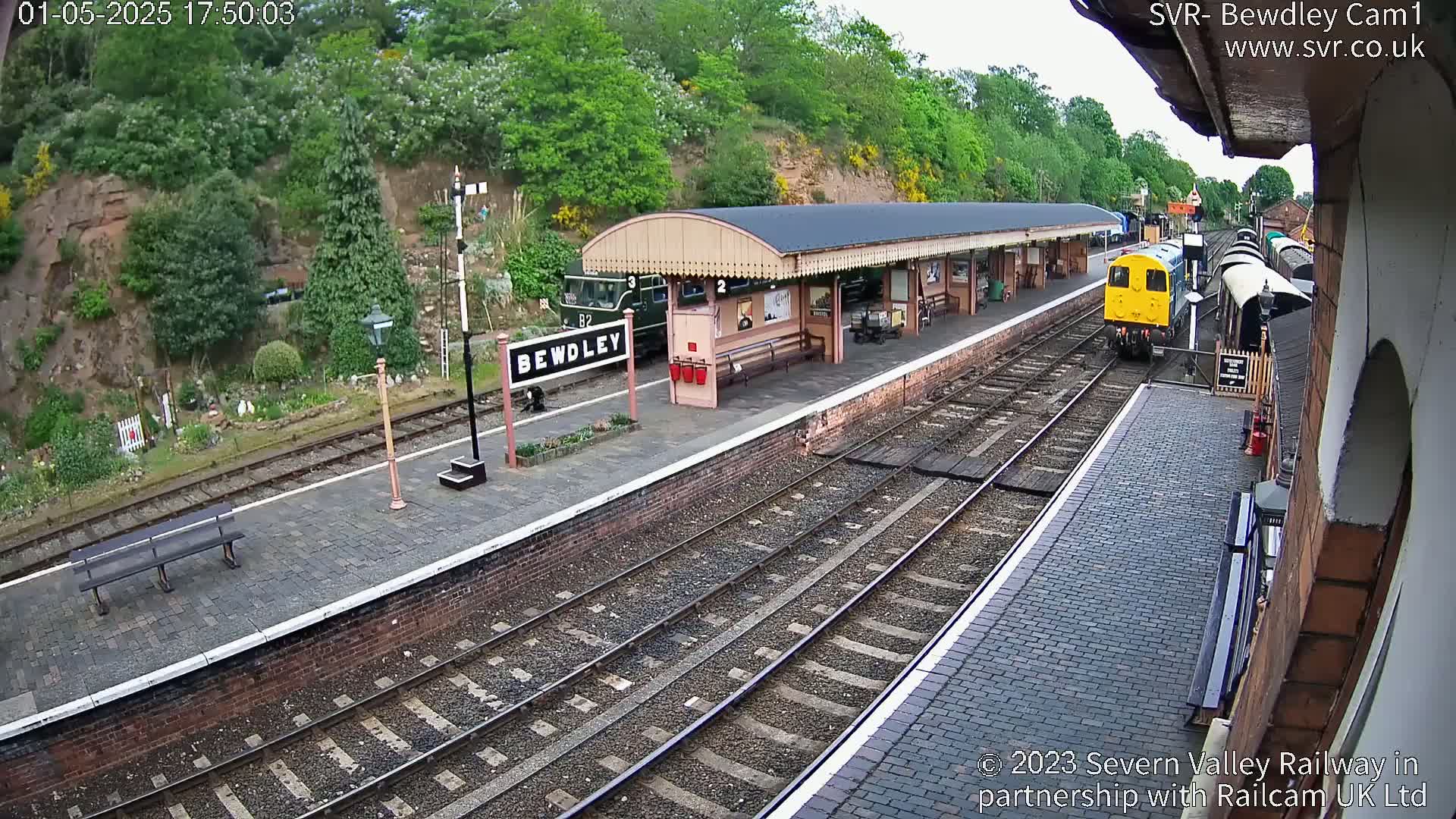 A train pulls into a station platform on a partly sunny day, with greenery visible beyond the tracks.