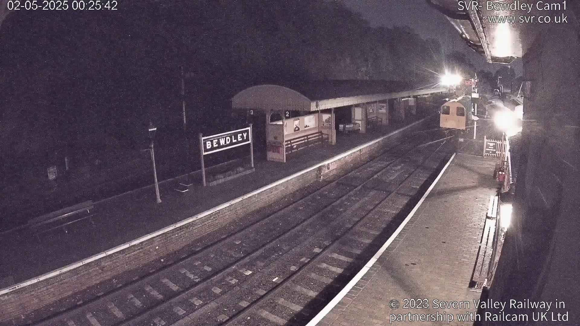 A nighttime view of a train station platform with a train partially visible, under dark conditions.