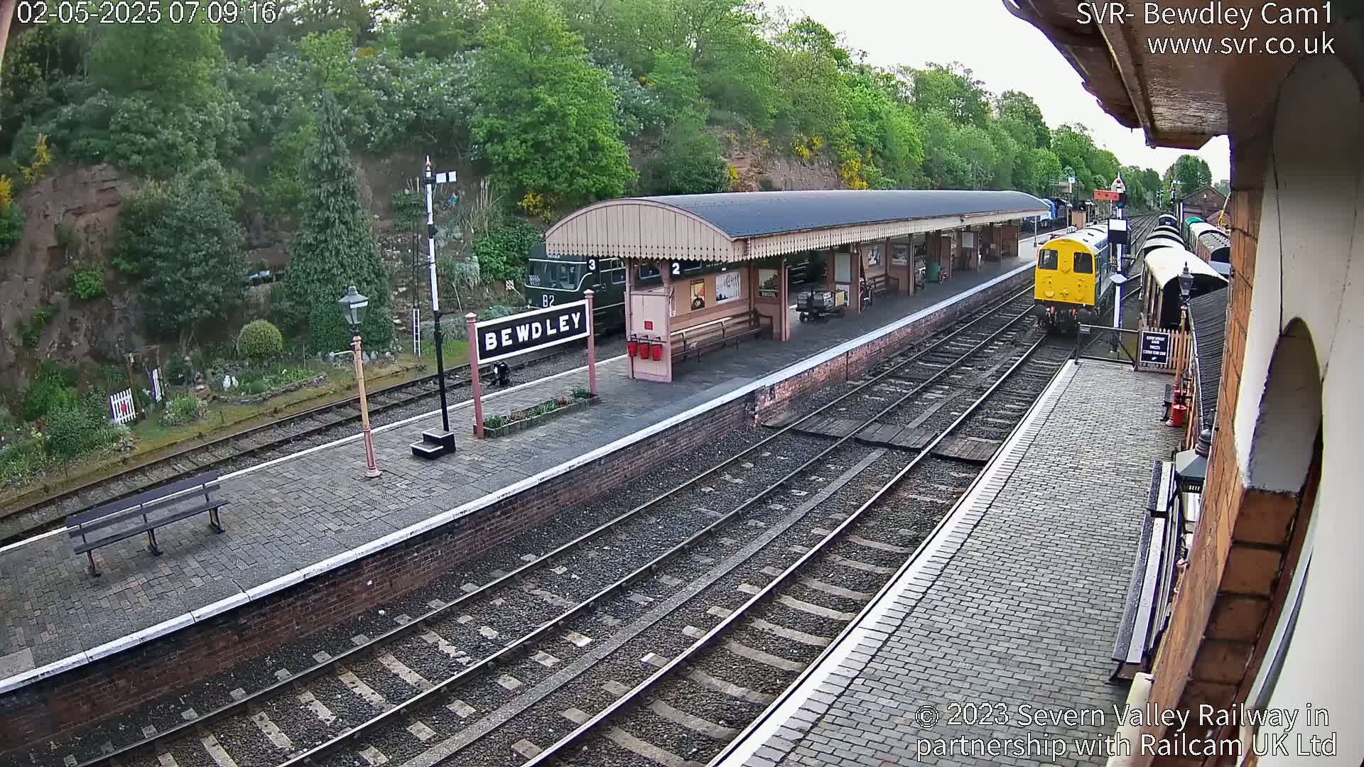 A yellow diesel train approaches a train station platform on a partly cloudy day, with a green passenger train visible further down the platform.