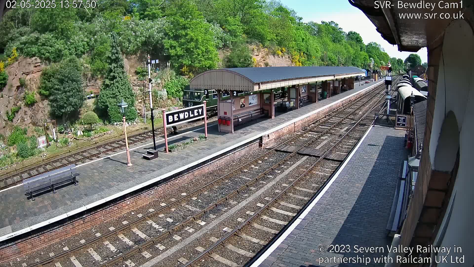 A train station platform with a train partially visible, situated next to a grassy hillside under sunny skies.