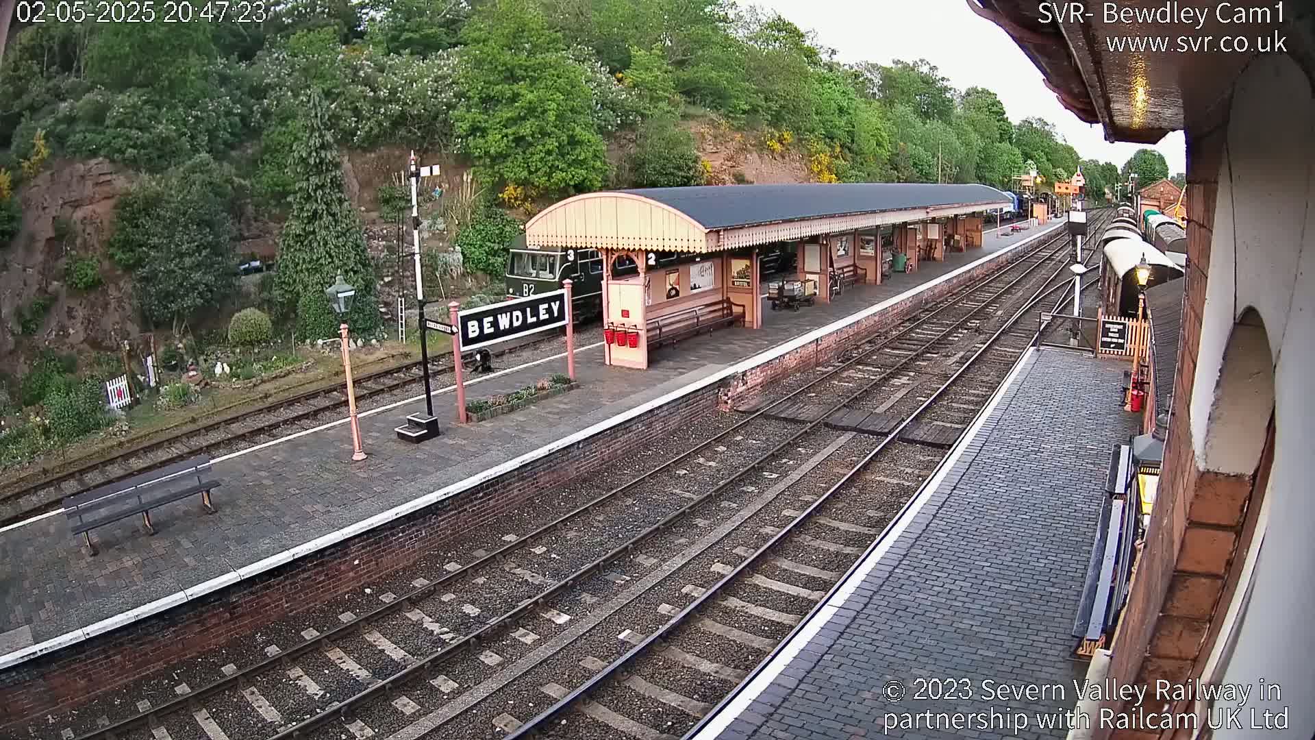 A train sits at a railway station platform under a covered waiting area, on an overcast evening.