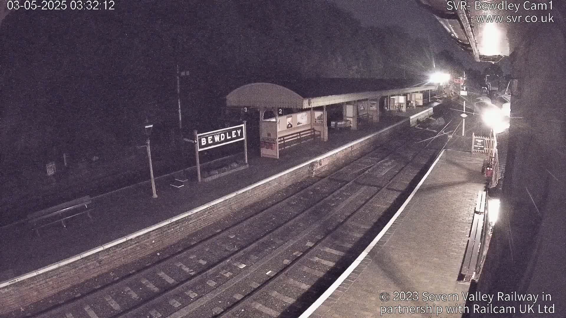 A nighttime view of a train station platform with train tracks and a waiting shelter under dim lighting.