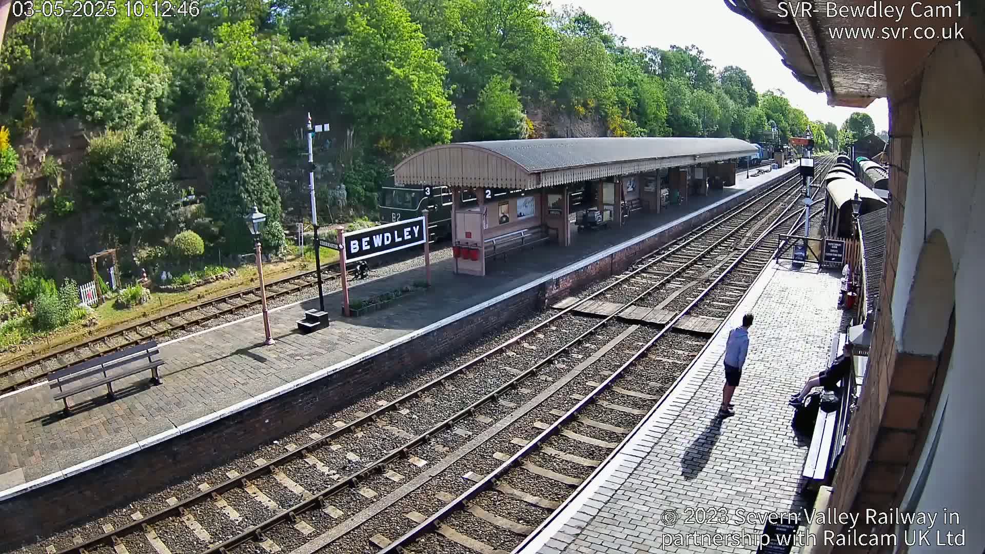 A sunny view of a train station platform with a covered waiting area, railway tracks, and a few people.