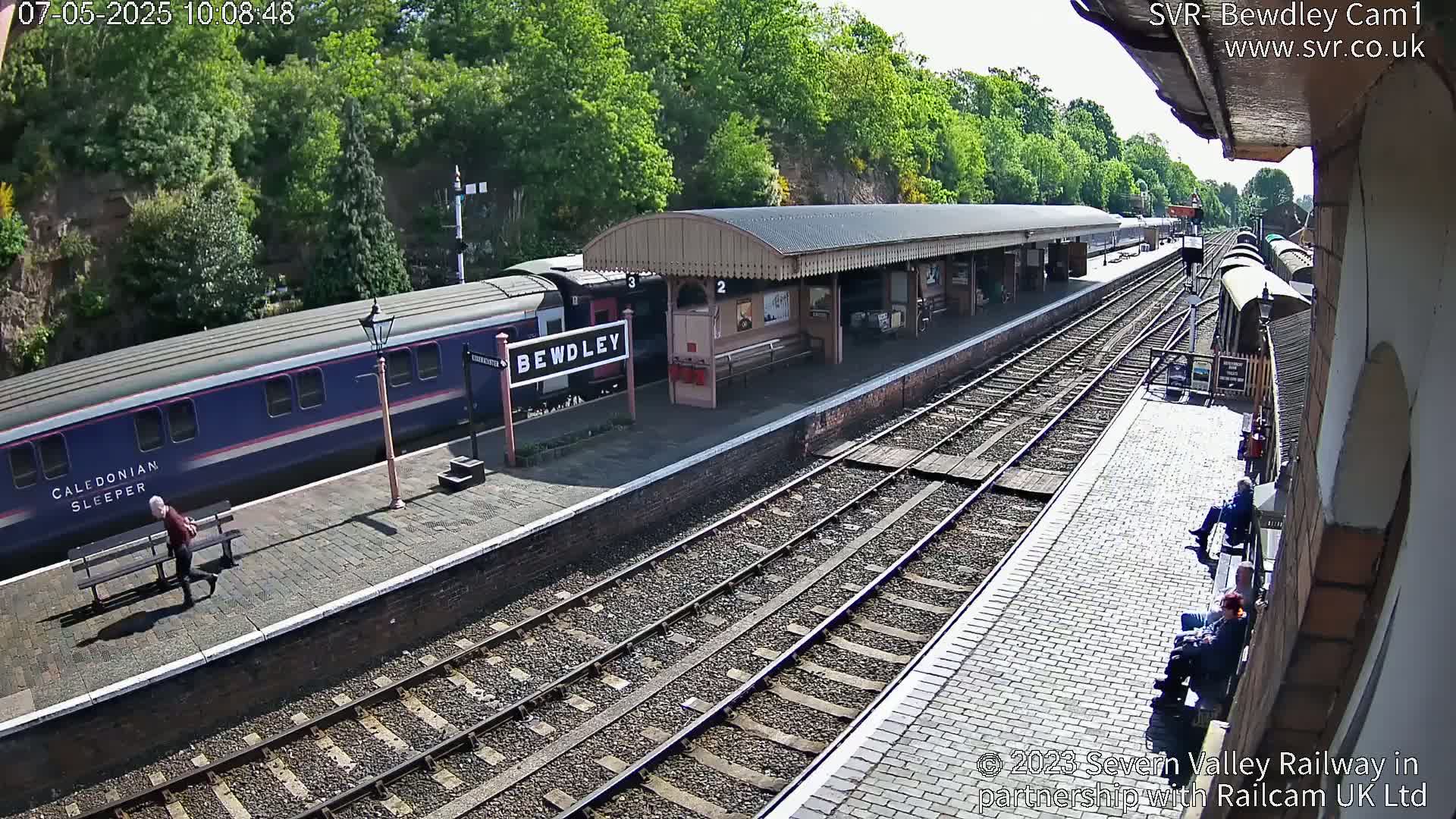 A long blue train is at a station platform on a sunny day, with several people near the tracks and a covered waiting area.