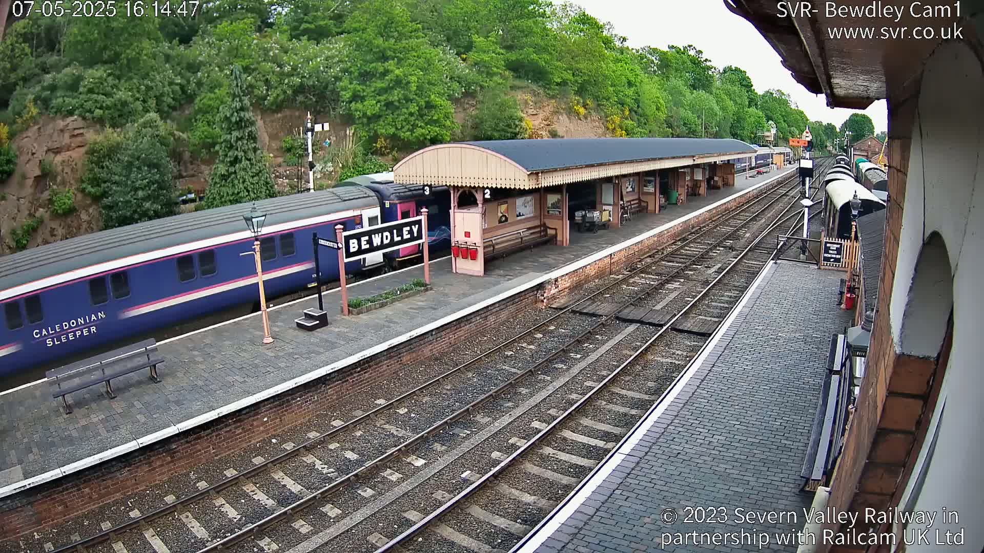 A blue train is stopped at a train station platform next to a covered waiting area, under a partly cloudy sky.