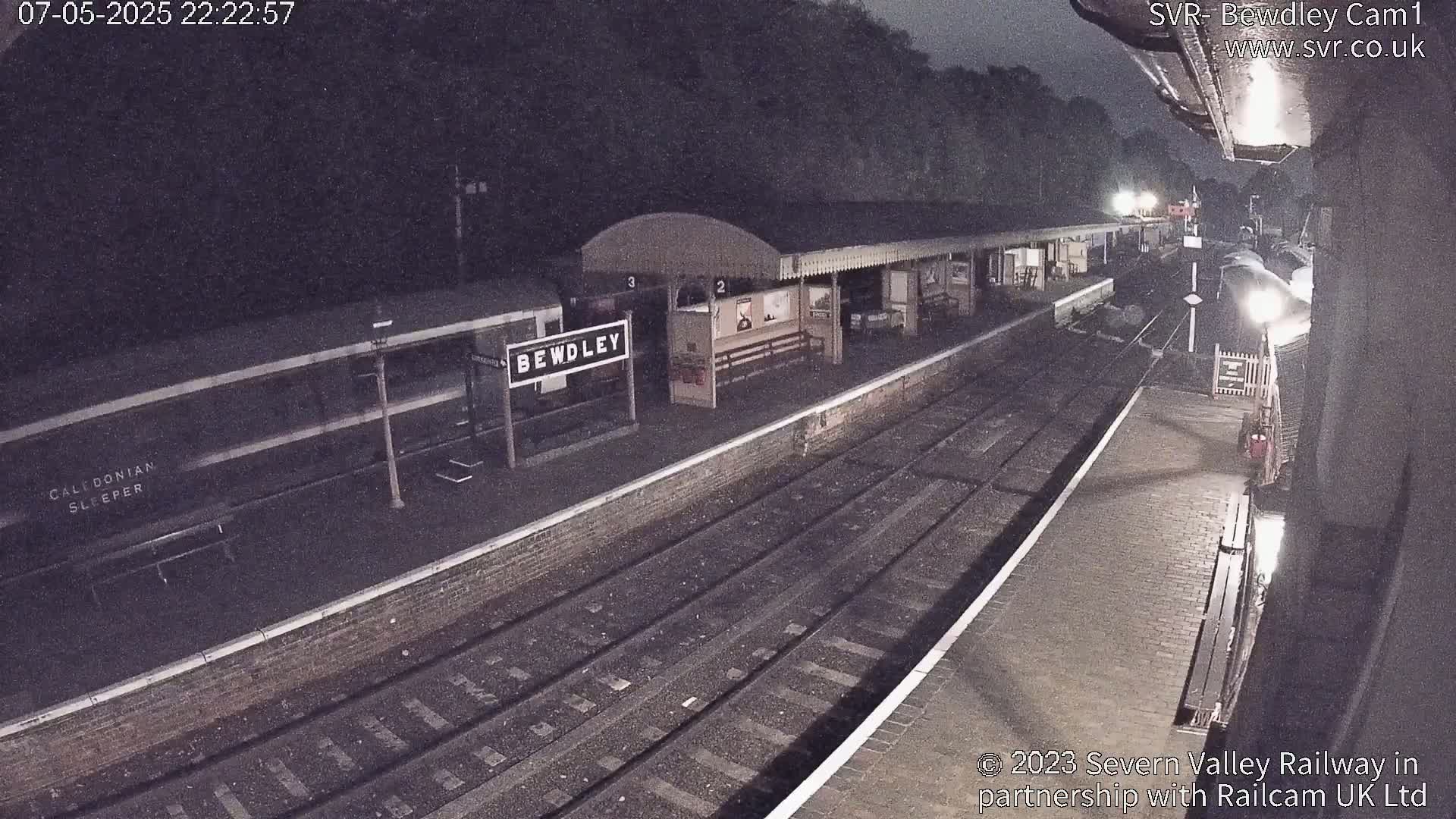A nighttime view of a train station platform with a train car visible, under dark skies.