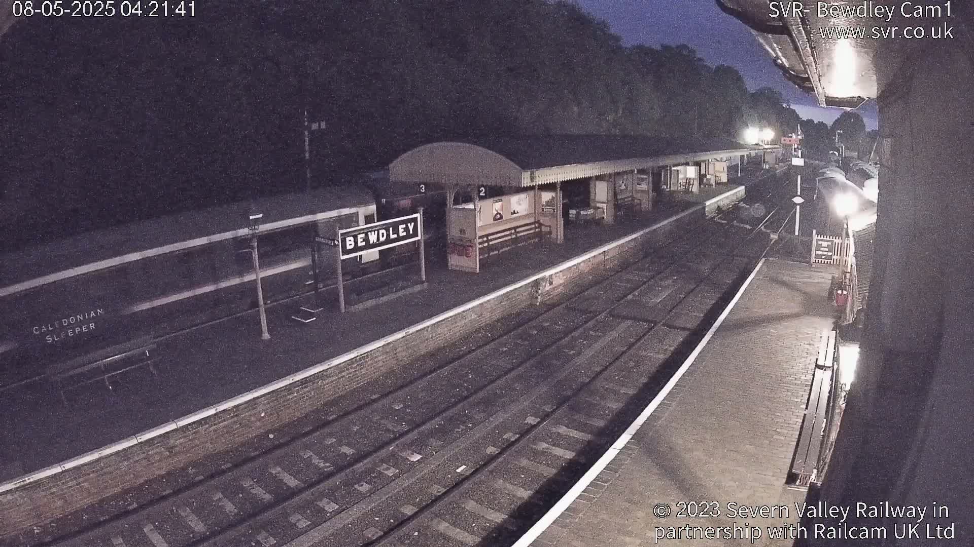 A nighttime view of a train station platform with a train and tracks, under dark skies.