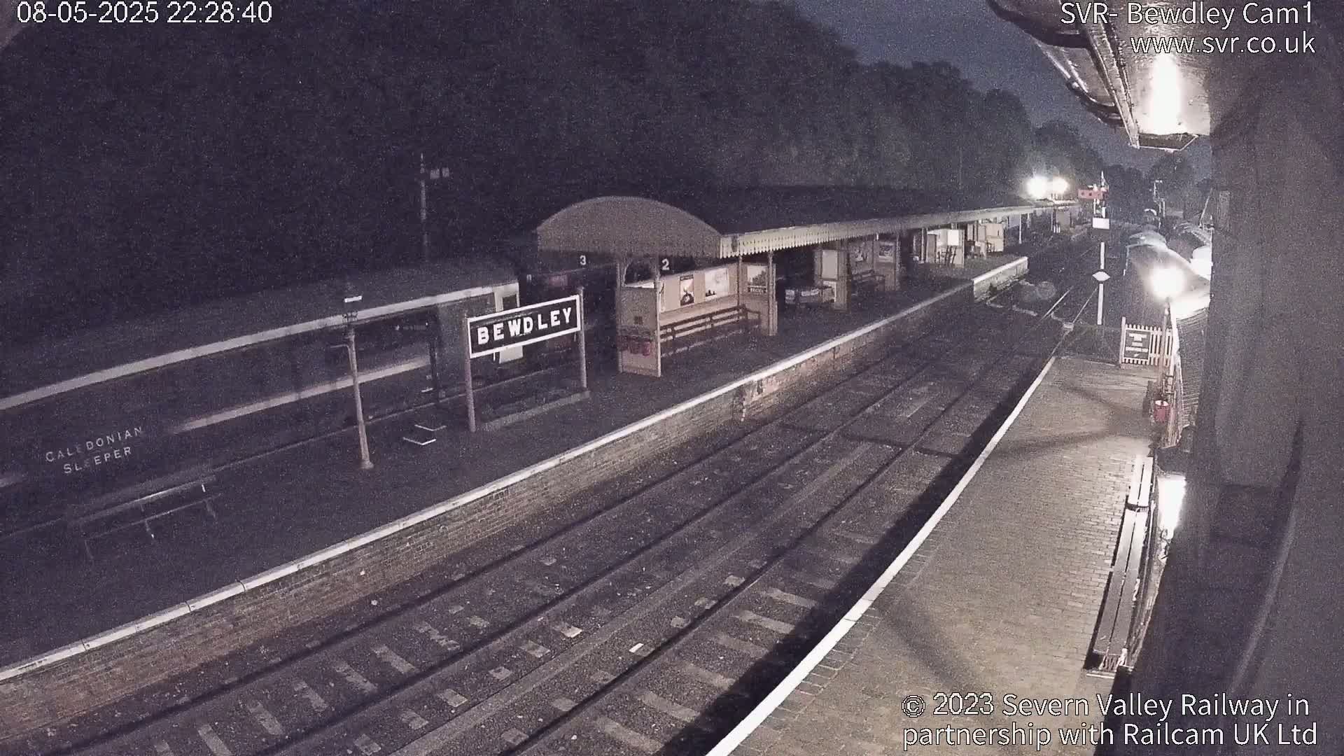 A nighttime view of a train station platform with a train, showing a mostly dark scene with some lights illuminating the platform and tracks.