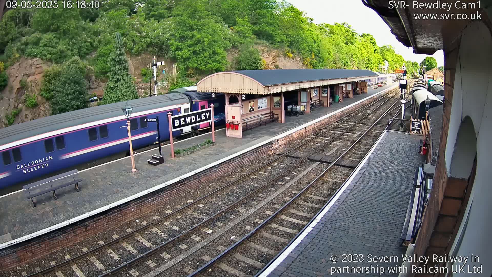 A blue and pink passenger train is at a station platform under a partly sunny sky; additional train cars are visible further down the tracks.
