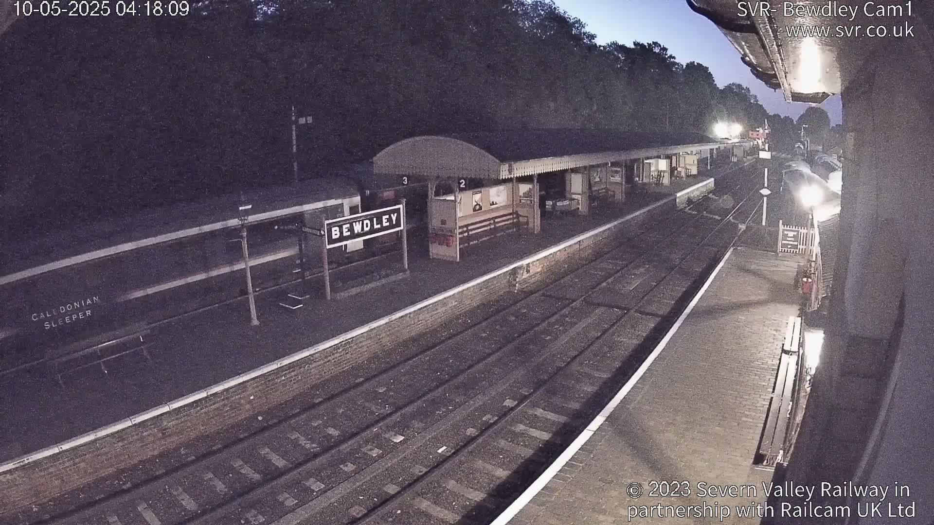 A train station at night, with a train partially visible and a platform under a covered awning, in low-light conditions.