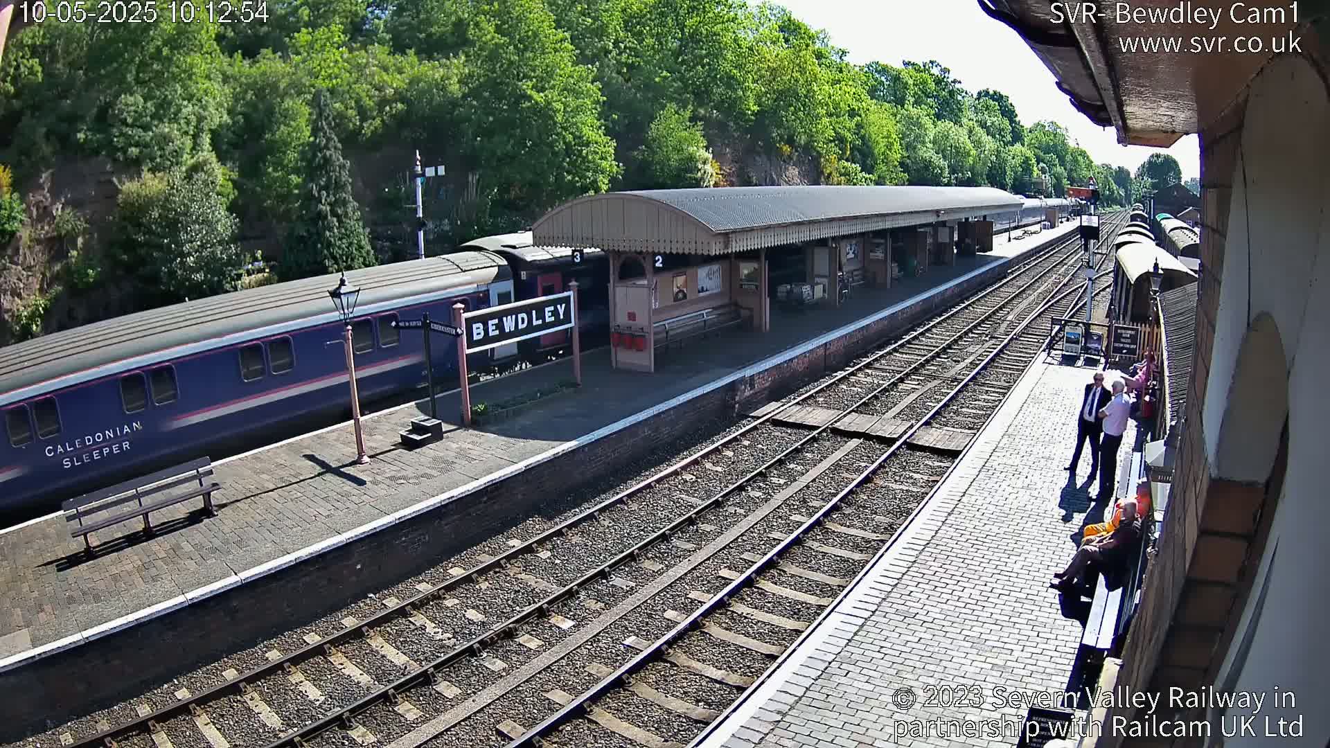 A blue and white passenger train is at a station platform on a sunny day, with several people standing and sitting nearby.