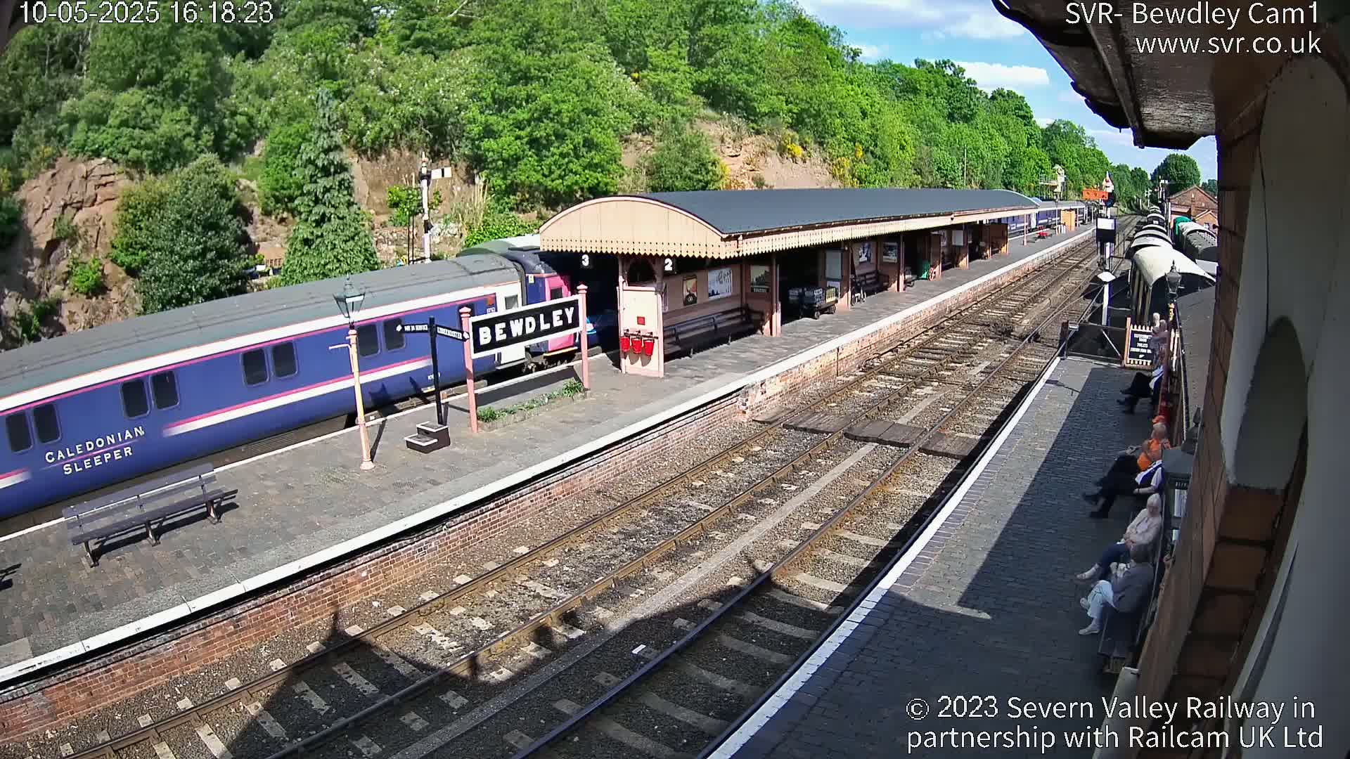 A blue and pink train is at a station platform on a sunny day, with several people sitting on benches nearby.