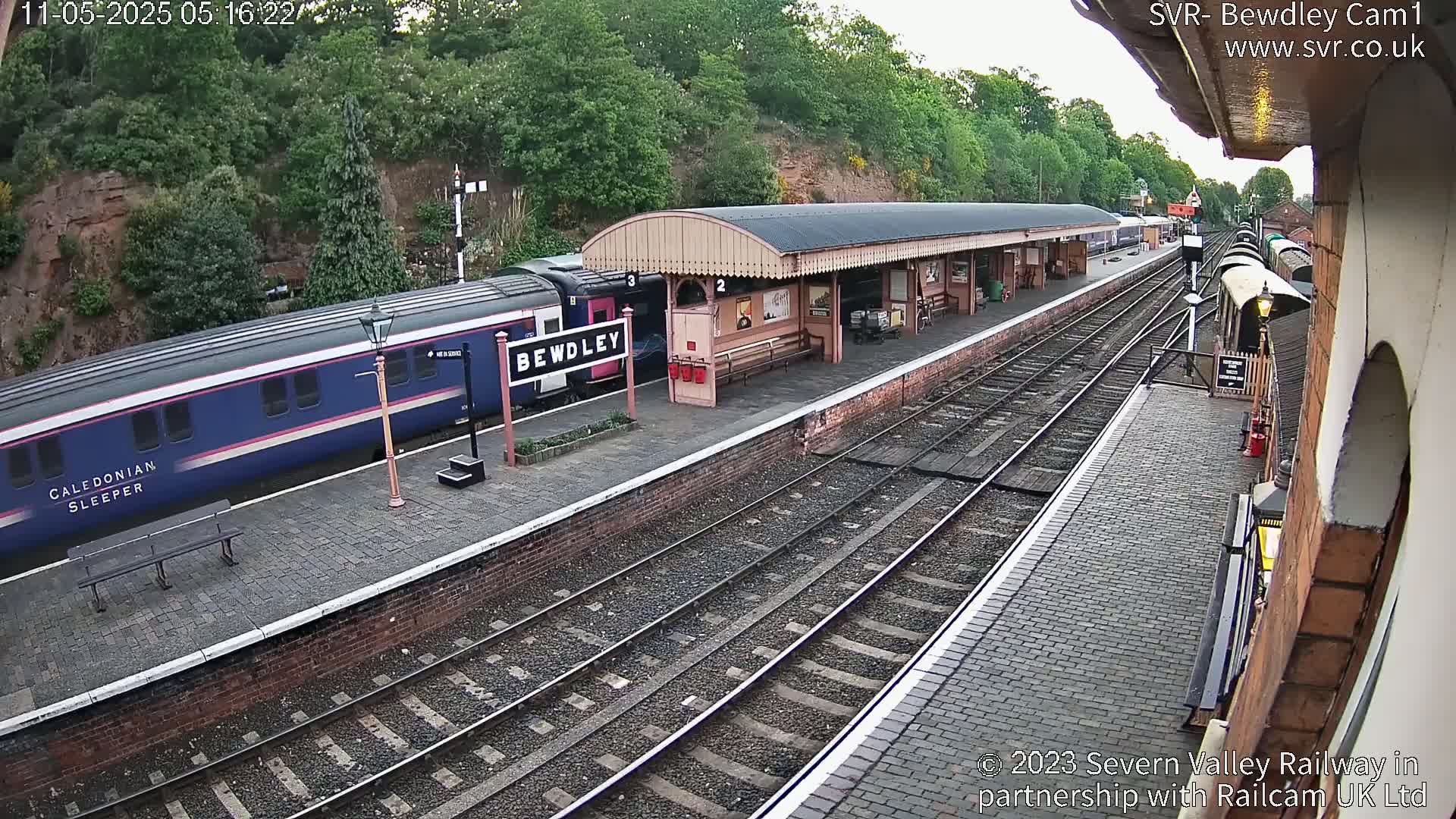 A blue Caledonian Sleeper train is at a station platform under an overcast sky, next to a covered waiting area.