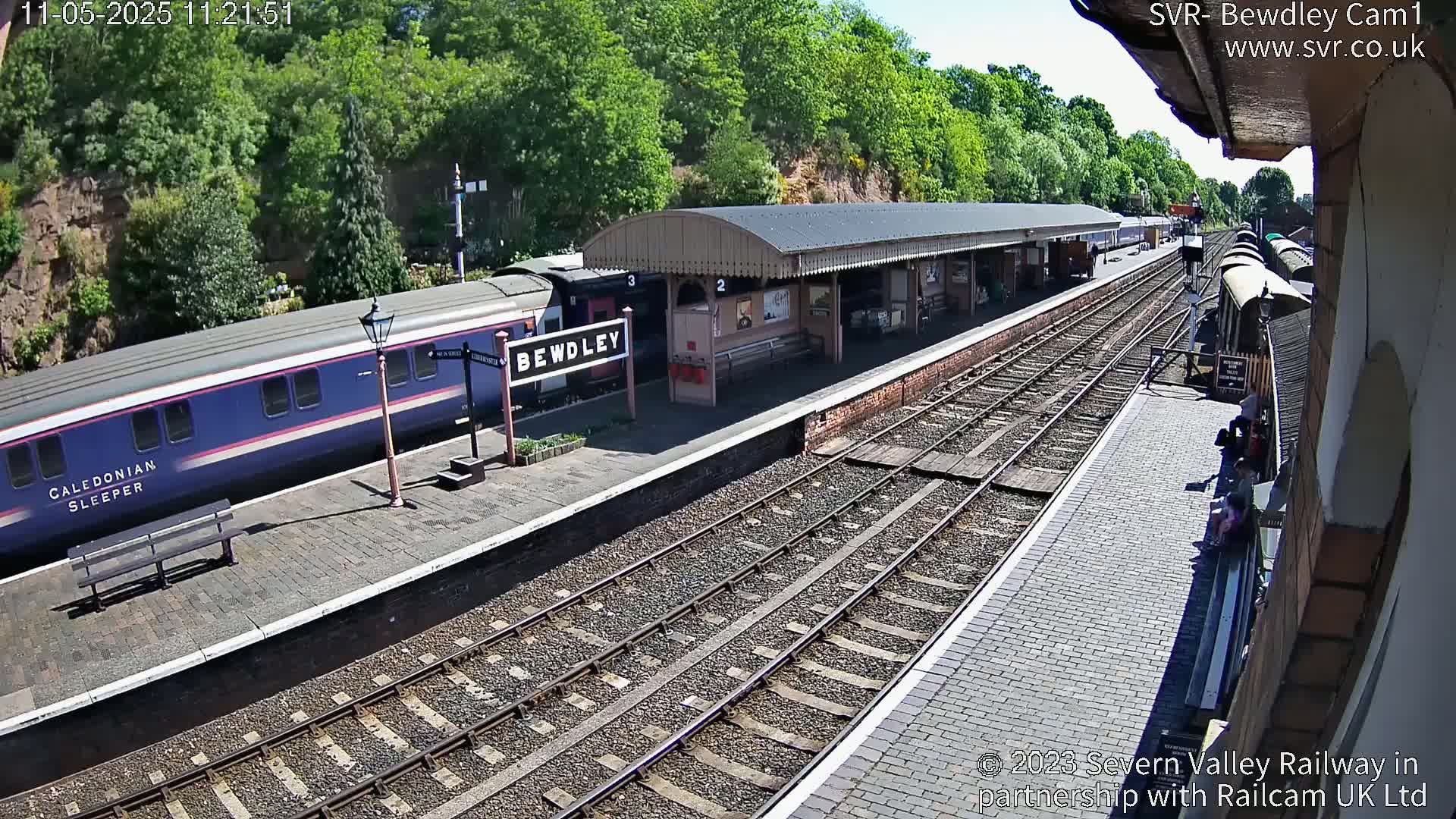 A blue and pink passenger train is stopped at a station platform on a sunny day, with other train cars visible further down the tracks and several people sitting on a bench.