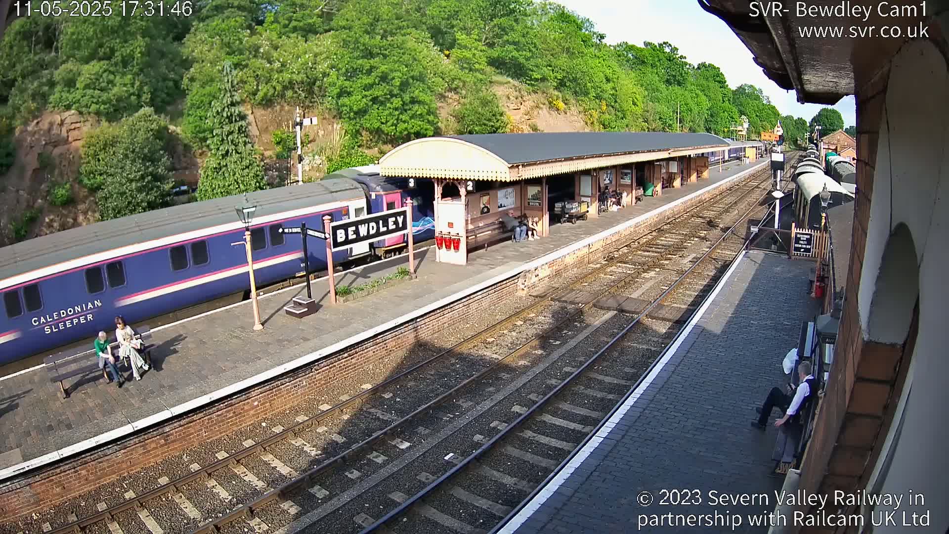A blue train is at a station platform on a sunny day, with a few people waiting on the platform.