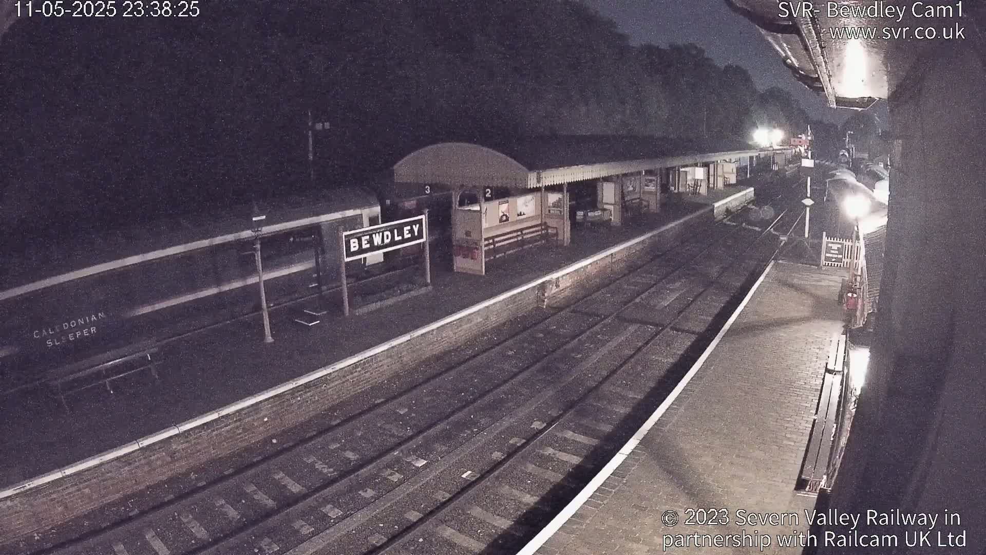 A nighttime view of a train station platform with a train alongside, under dark skies.