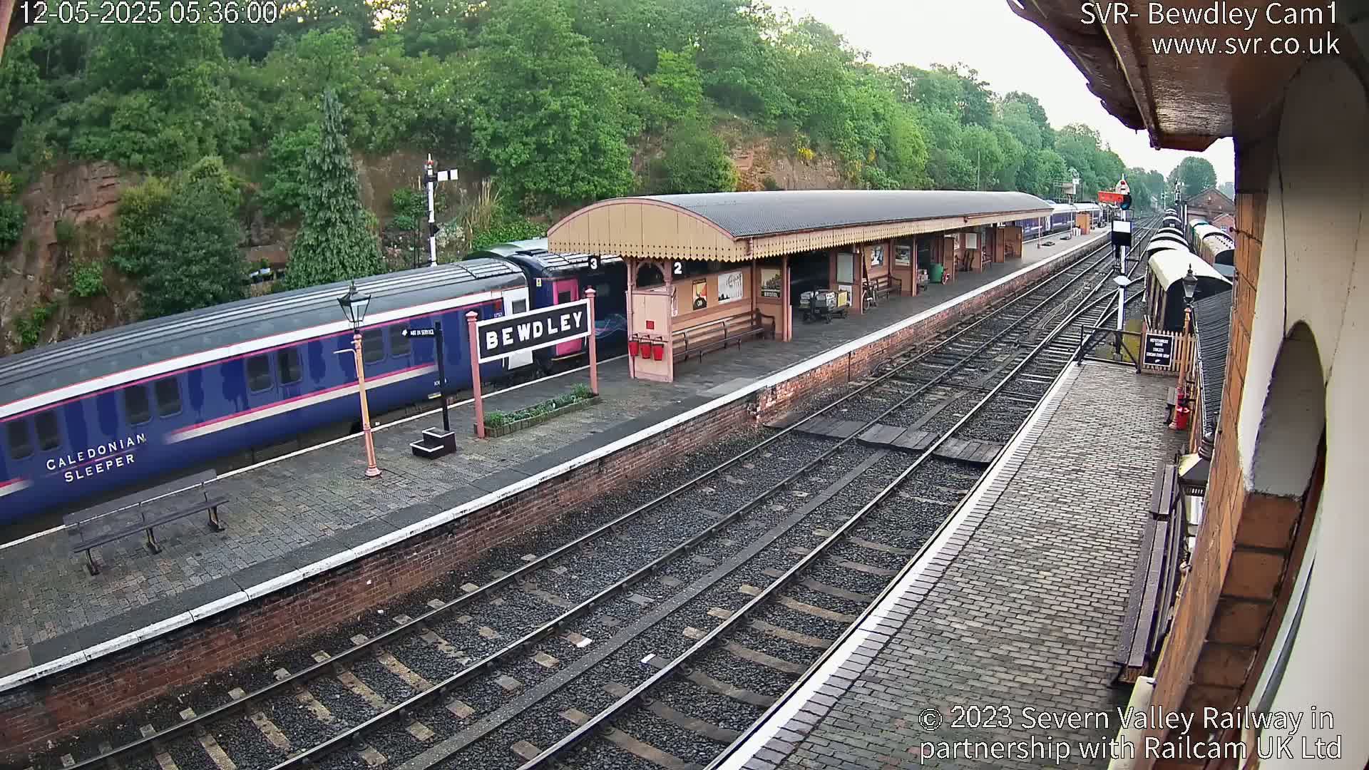 A blue and pink Caledonian Sleeper train is at a station platform on a partly cloudy day, with multiple train tracks visible.