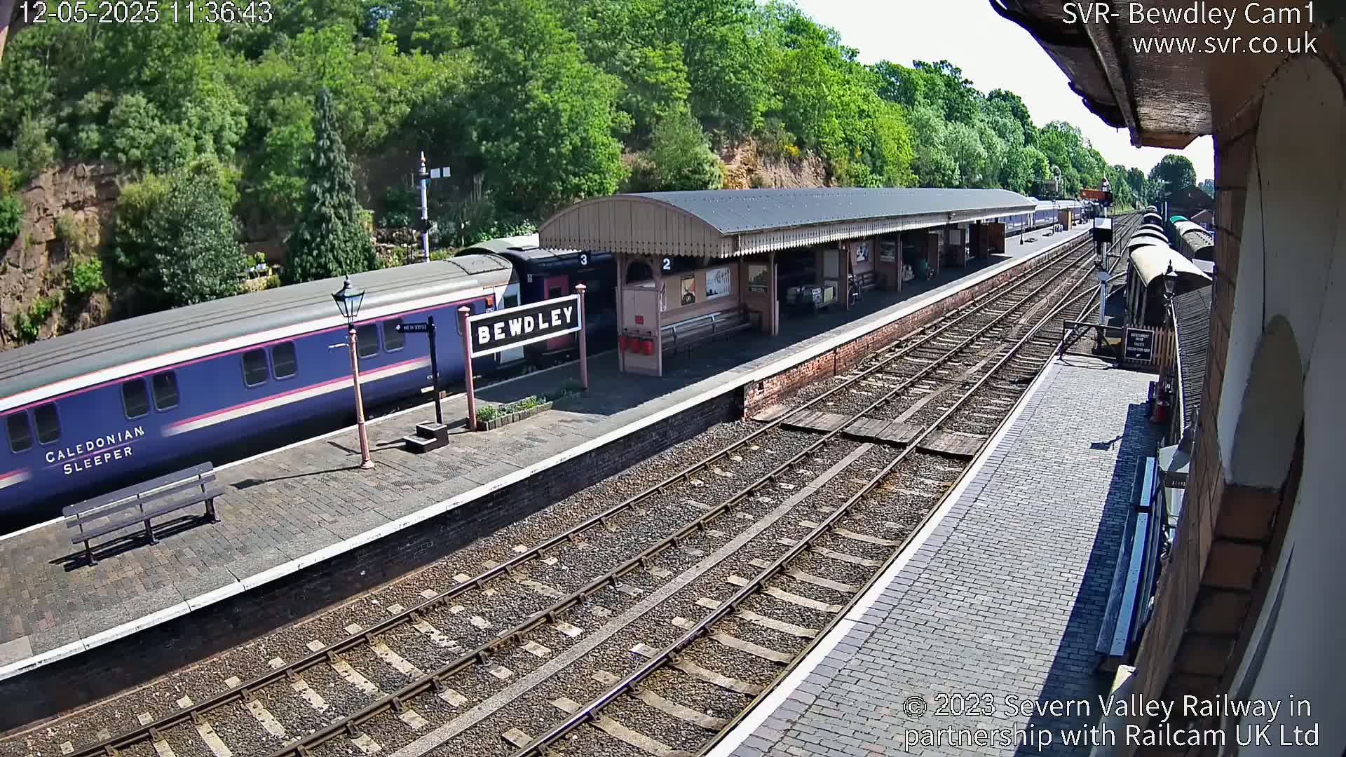 A long blue train is at a station platform on a sunny day, next to train tracks extending into the distance.