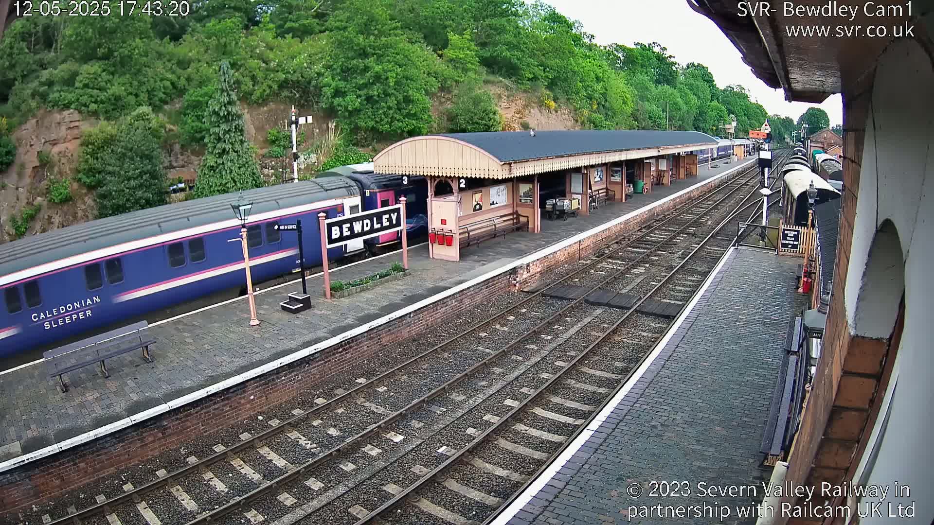 A blue and pink Caledonian Sleeper train is at a station platform next to a covered waiting area on a partly sunny day.