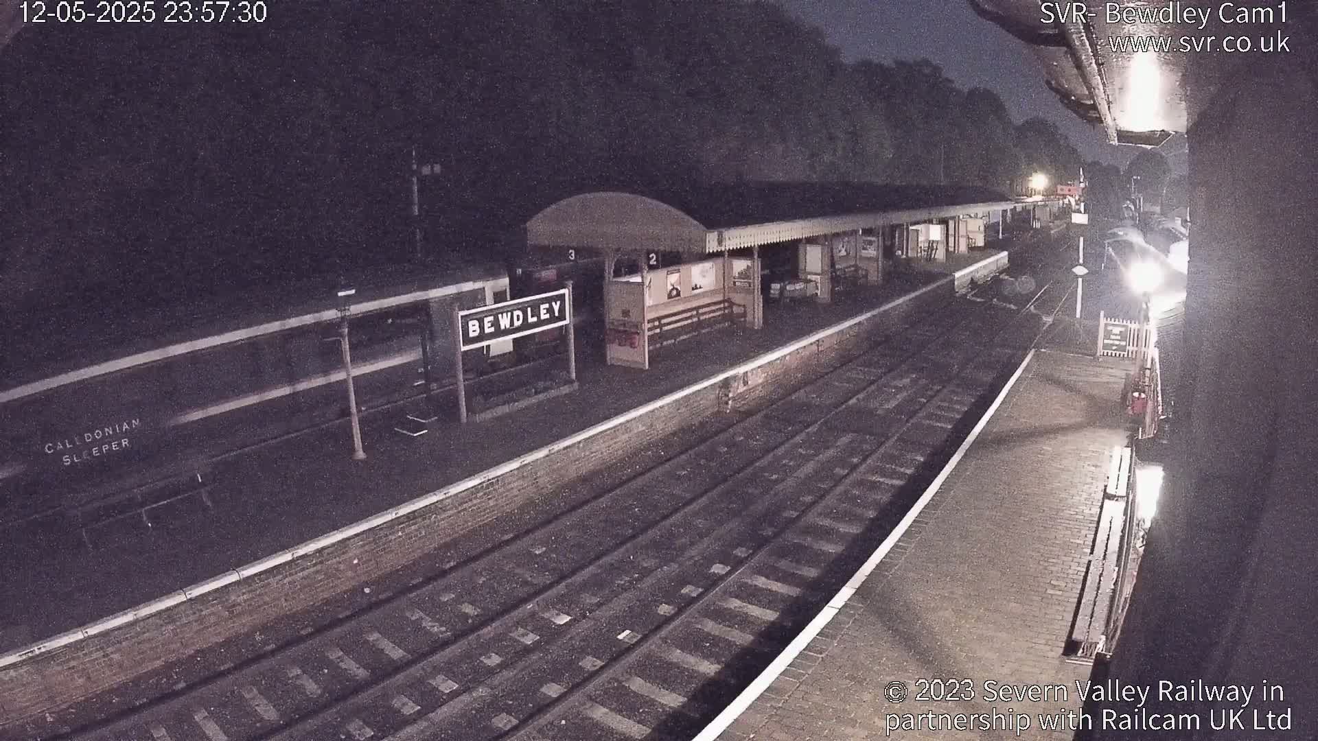 A nighttime view of a train station platform with a train and a station building, under dark skies.