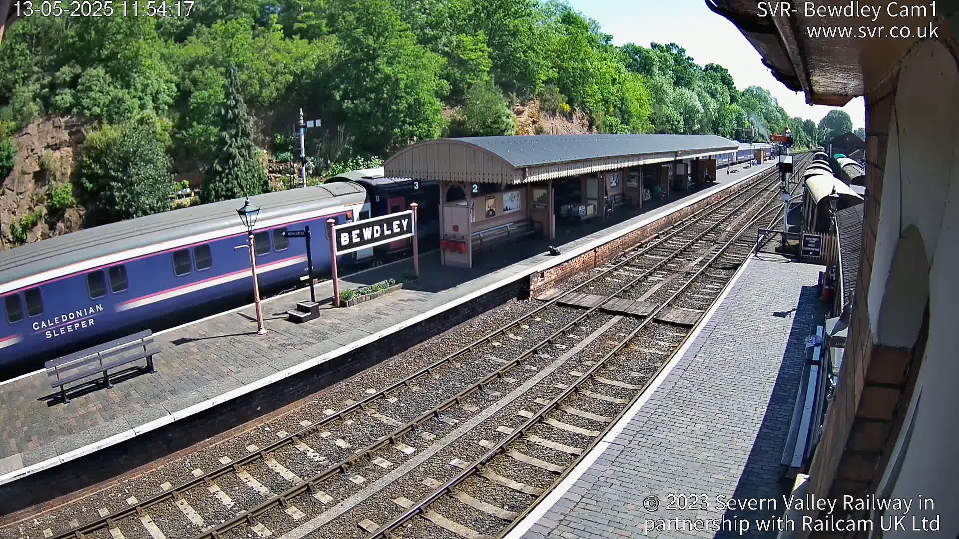 A blue passenger train is stopped at a train station platform on a sunny day, with additional train cars visible further down the tracks.