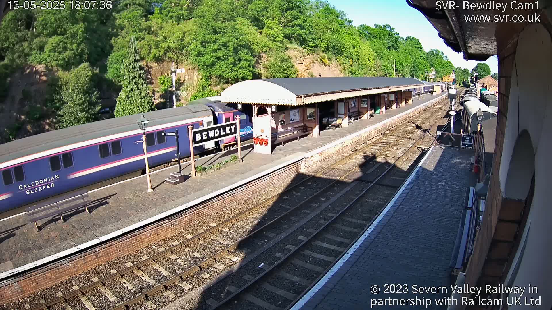 A blue Caledonian Sleeper train is at a station platform on a sunny day.