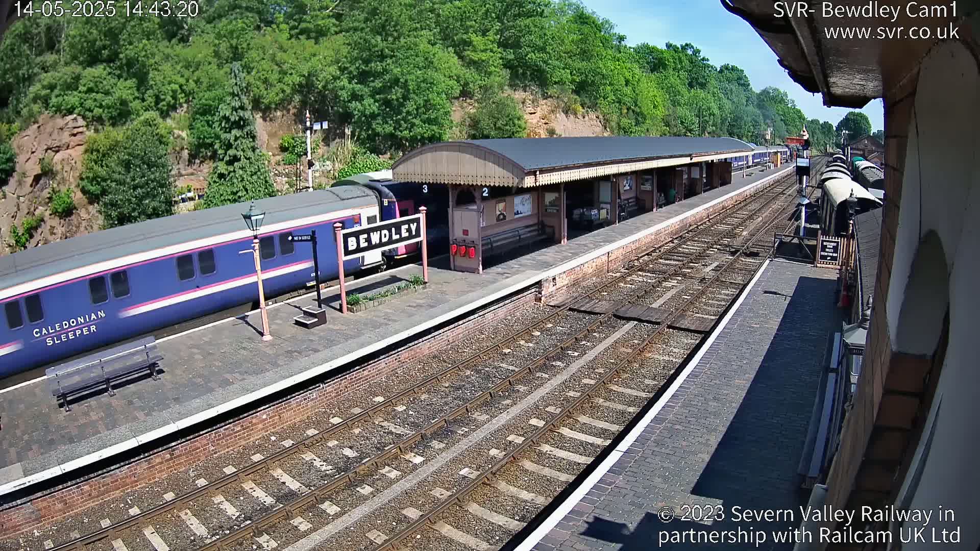 A blue passenger train is at a railway station platform on a sunny day, with additional rail cars visible further down the tracks.