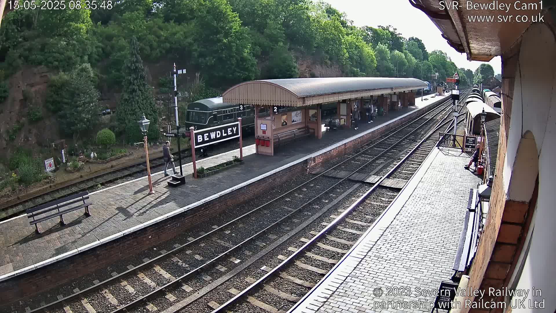 A sunny view of a train station platform with a dark green passenger train and several people visible, and a brick wall and several train tracks.