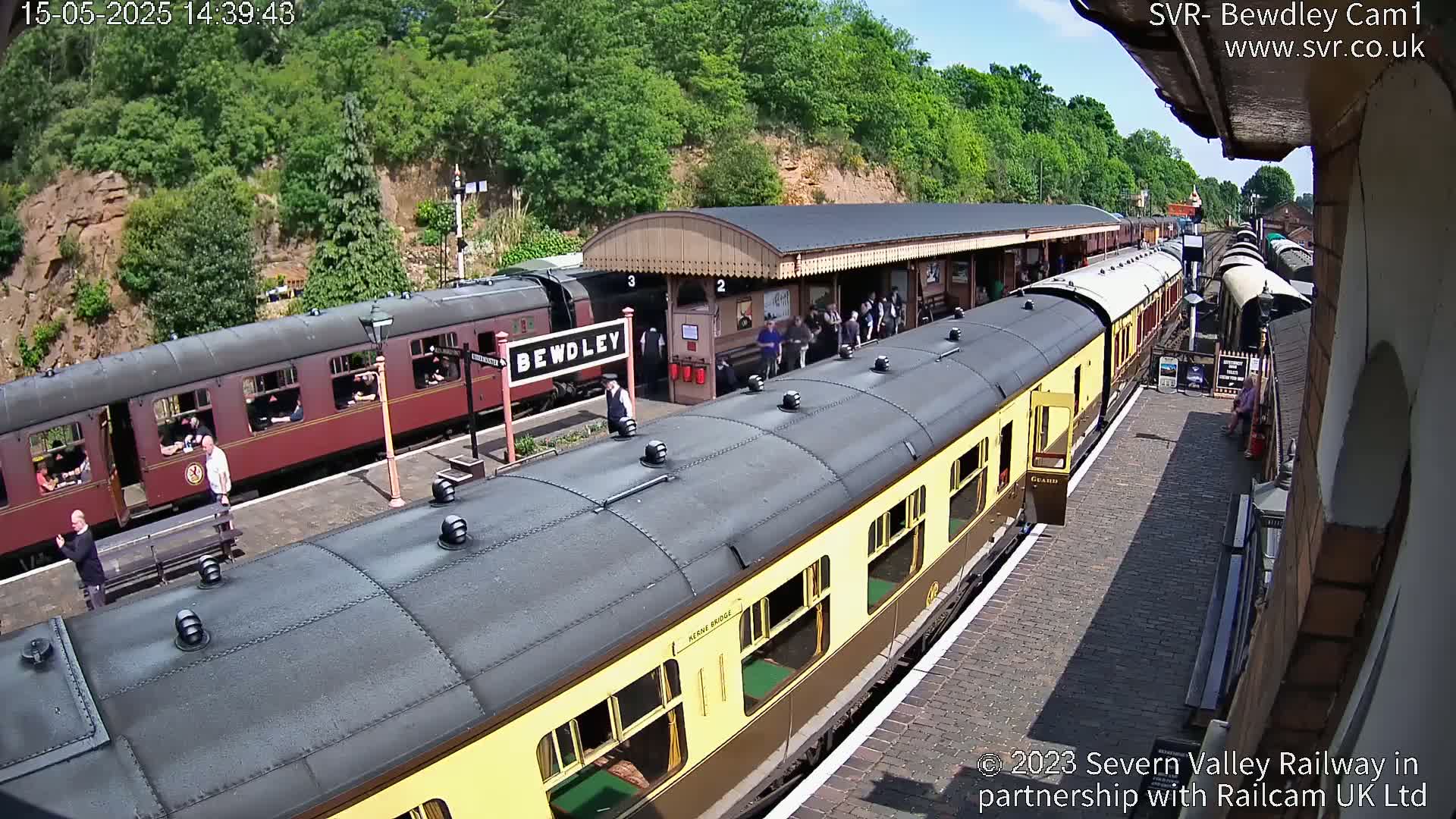 A sunny day at a train station shows two vintage passenger trains at a platform with several people standing and sitting nearby.