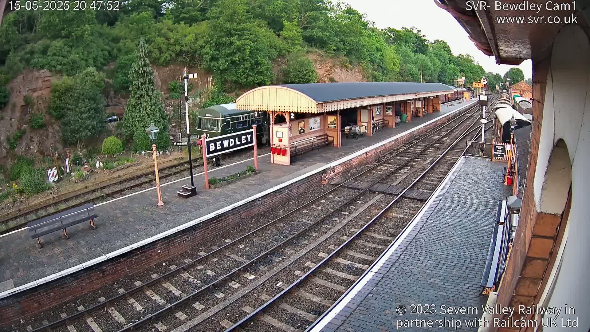 A train sits at a station platform next to a covered waiting area, under a partly cloudy sky.
